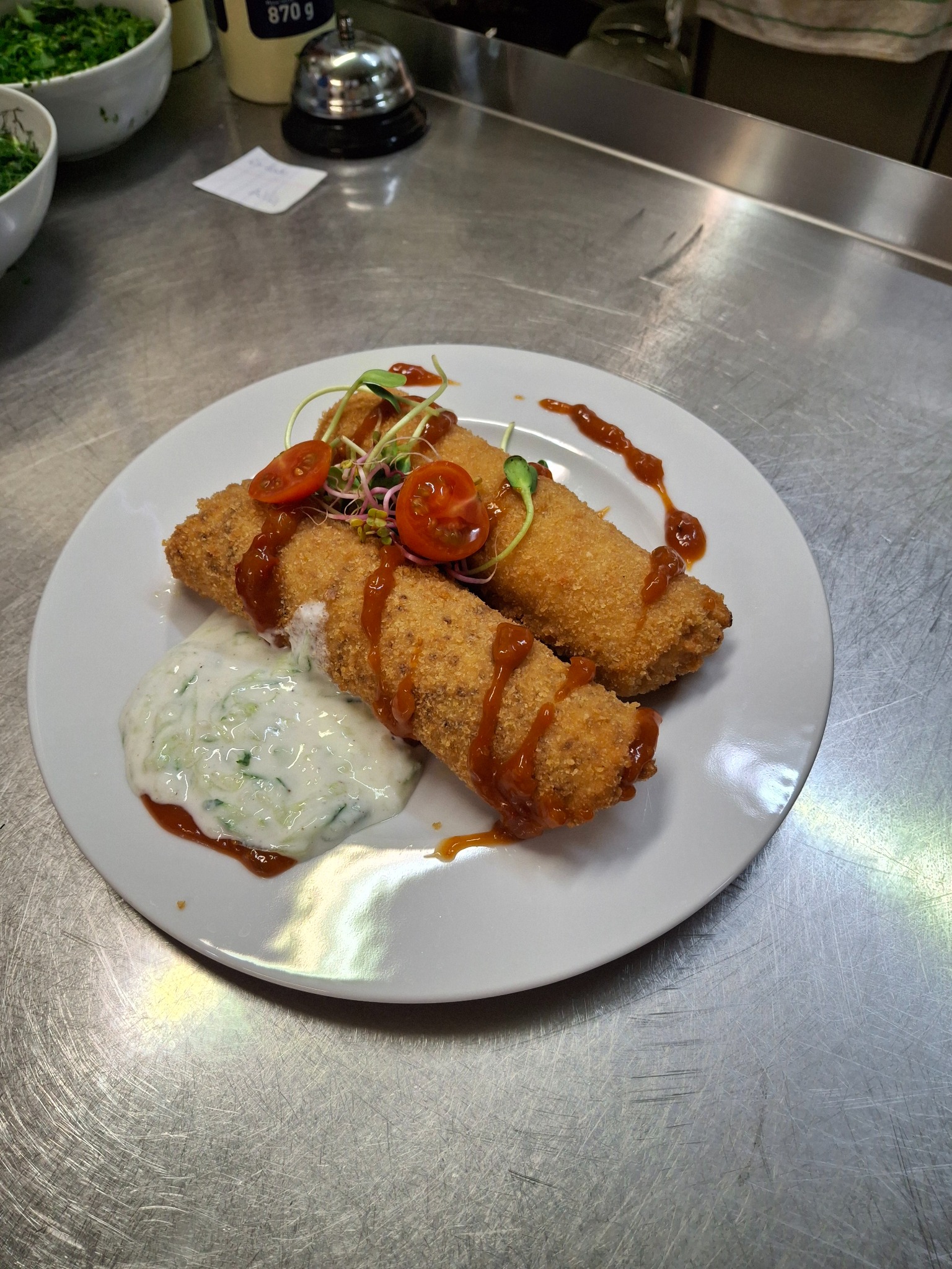 Dwa krokiety panierowane na białym talerzu, polane sosem, z dodatkiem dipu ogórkowego, udekorowane kiełkami i pomidorkami cherry, na tle metalowego blatu.