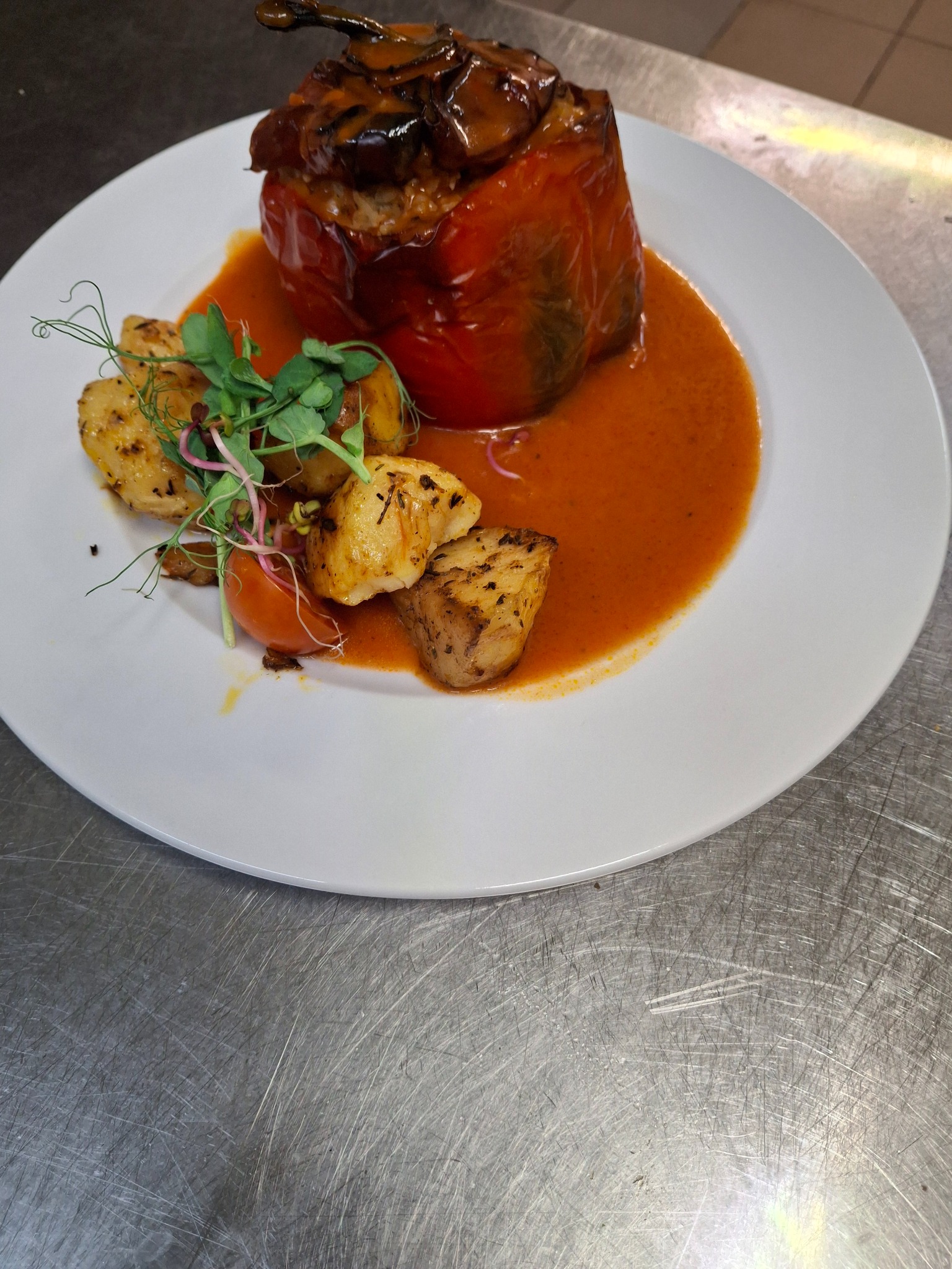 Czerwona papryka faszerowana na białym talerzu, polana sosem pomidorowym, z dodatkiem pieczonych ziemniaków i świeżych kiełków, na tle metalowego blatu.