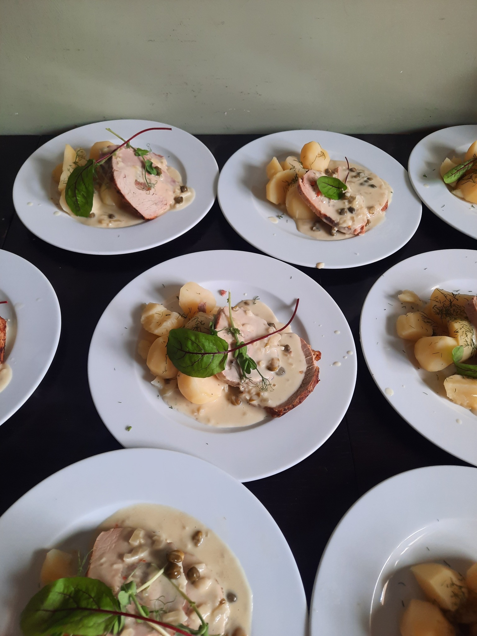 Ułożone rzędem talerze z porcjami pieczonego mięsa w kremowym sosie kaparowym, podanego z gotowanymi ziemniakami i dekoracją z ziół.