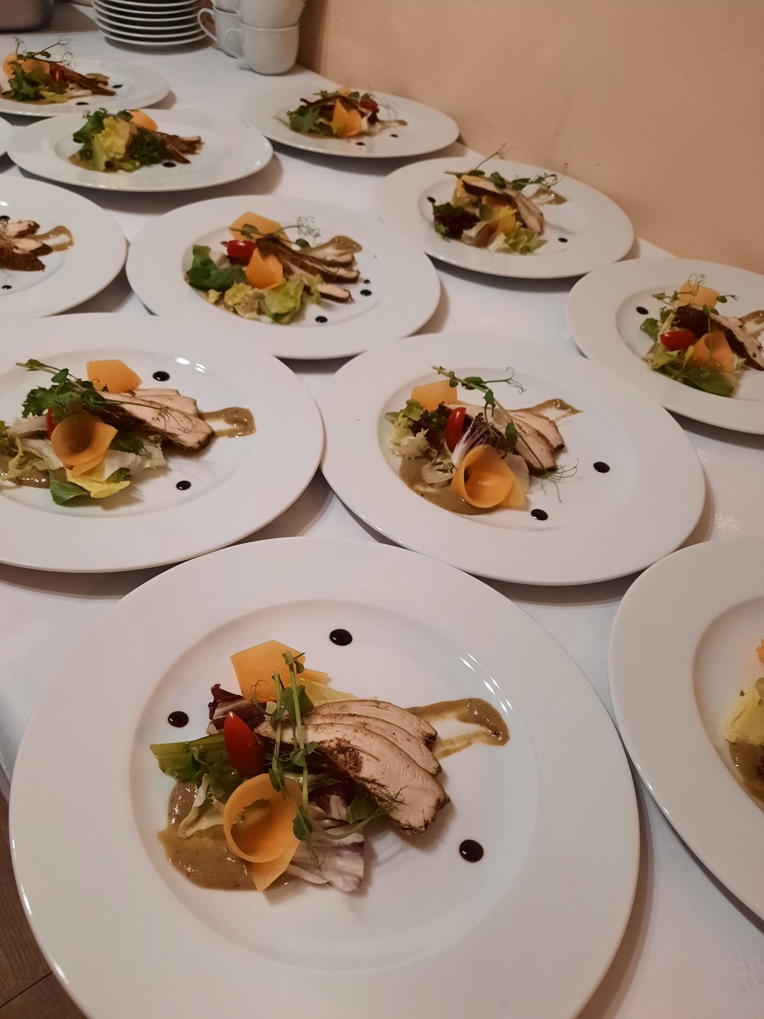 Ułożone rzędem talerze z sałatką z grillowanym kurczakiem, melonem, pomidorem i sosem balsamicznym, przygotowane na białym obrusie, widoczne filiżanki w tle.