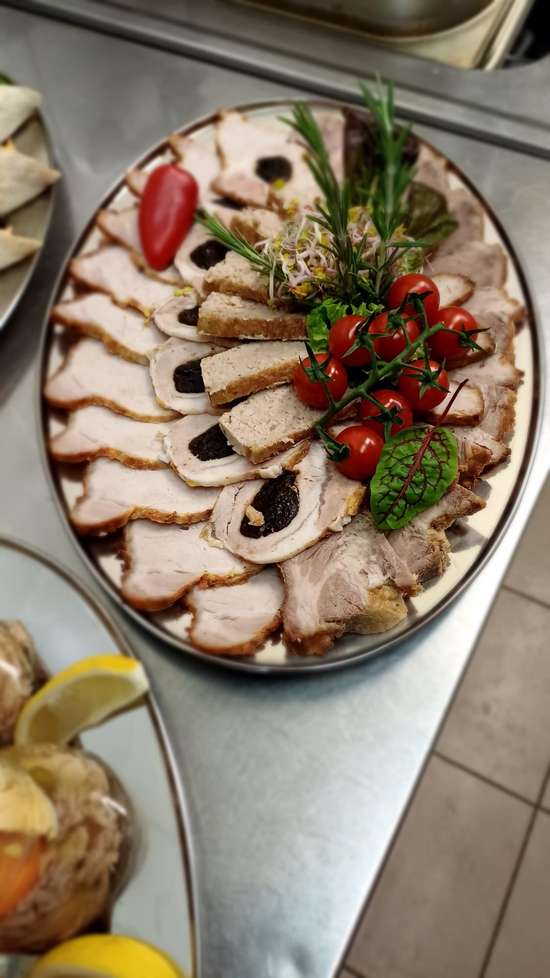 Owalny półmisek z plastrami pieczonego mięsa z suszoną śliwką, pasztetu, dekorowany gałązkami rozmarynu, kiełkami i pomidorkami cherry na blacie ze stali nierdzewnej.