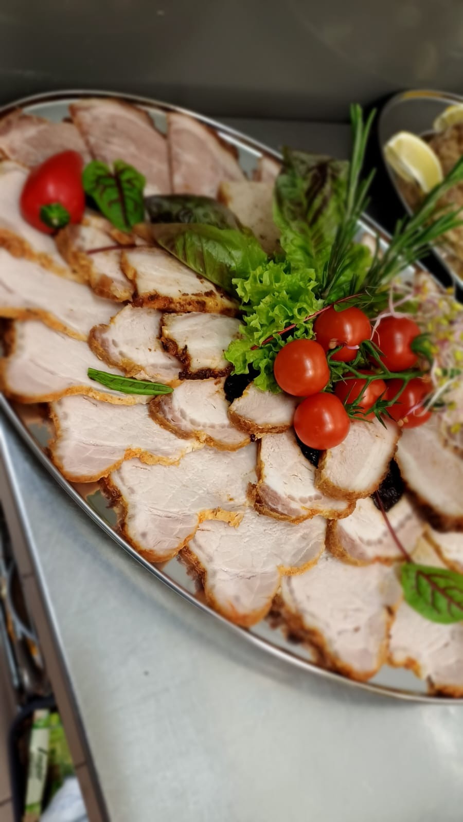 Srebrna taca z plastrami pieczonego mięsa, udekorowana pomidorkami cherry, gałązkami rozmarynu, sałatą i kiełkami, z perspektywy lekko z góry.