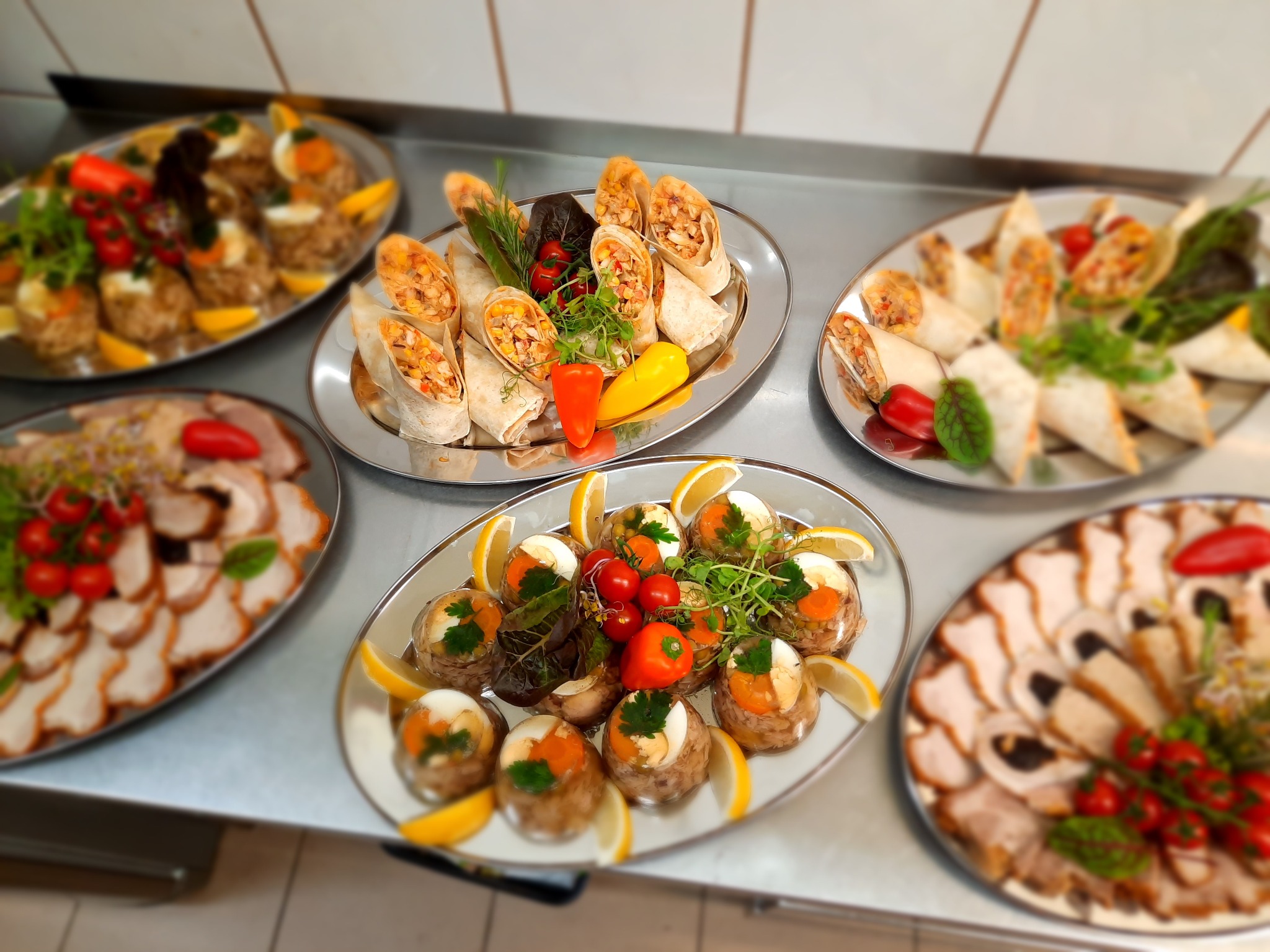 Srebrne tace z różnorodnymi przekąskami: roladki z tortilli z warzywami i kurczakiem, galaretki z jajkiem i warzywami, plastry mięsa z dodatkami na metalowym stole.