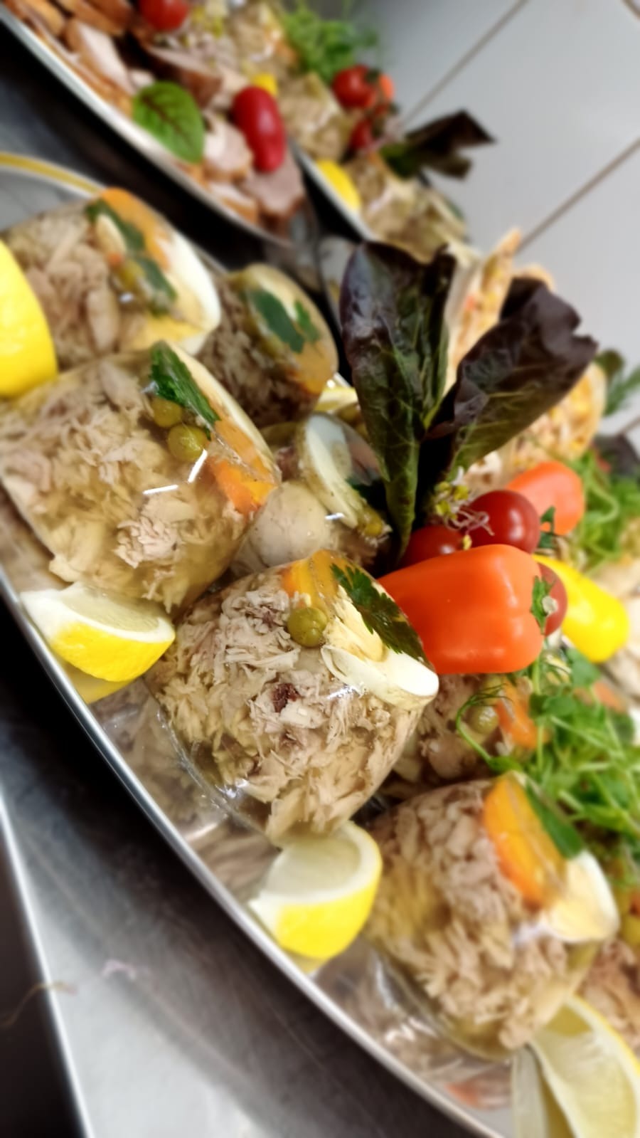 Galaretki z mięsem i warzywami, udekorowane cytryną, papryką i zielonymi listkami na srebrnej tacy, prezentowane pod kątem.