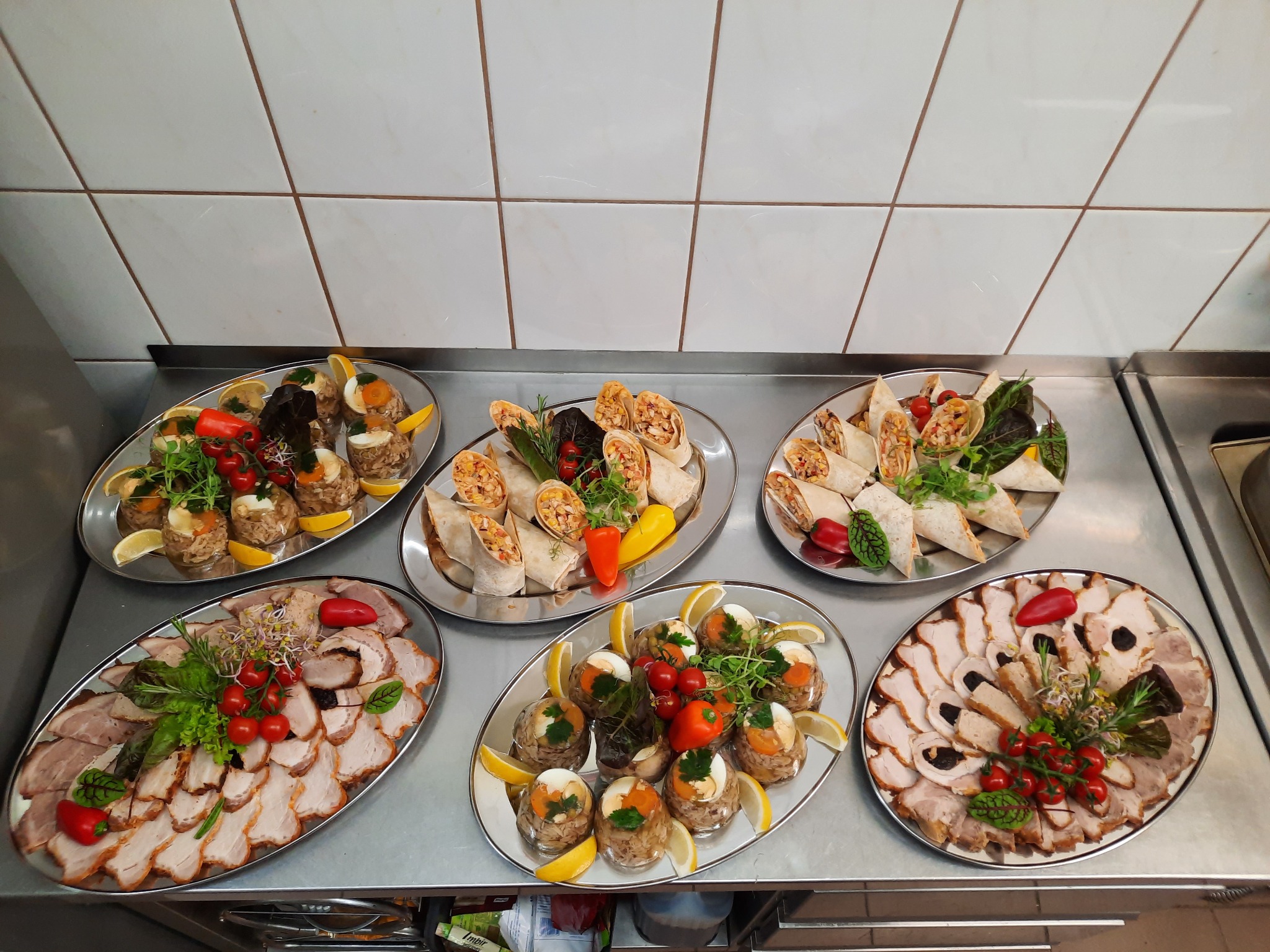 Sześć srebrnych pater z różnorodnymi przekąskami: roladki tortilli z warzywami, galaretki z jajkiem i mięsem, plastry pieczonego mięsa z dekoracjami z warzyw, ułożone na metalowym blacie w kuchni...