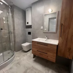 Wykończona łazienka z kabiną prysznicową, umywalką z drewnianą szafką i toaletą w odcieniach szarości z akcentami drewna. Grzejnik elektryczny na ścianie.