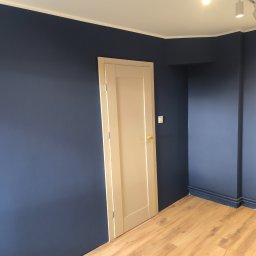 MAJSTER SKW-A - Pokój po malowaniu ścian na granatowo i ułożeniu paneli podłogowych. Widoczne białe drzwi i oświetlenie sufitowe.
