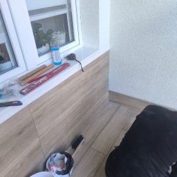 MAJSTER SKW-A - Układanie płytek imitujących drewno na podłodze i ścianie, tworzących boazerię. Pracownik klęczy, obok narzędzia i białe okno z rośliną.