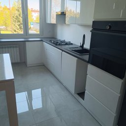 MAJSTER SKW-A - Nowoczesna kuchnia z białymi, lakierowanymi szafkami, ciemnym blatem, czarnym zlewem i płytą gazową, z wbudowanym piekarnikiem i jasnymi płytkami.