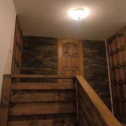 MAJSTER SKW-A - Drewniane schody z balustradą prowadzące do korytarza z ciemną, fakturowaną okładziną ścienną imitującą kamień. W centrum drewniane drzwi z napisem WC, oświetlone lampą sufitową.