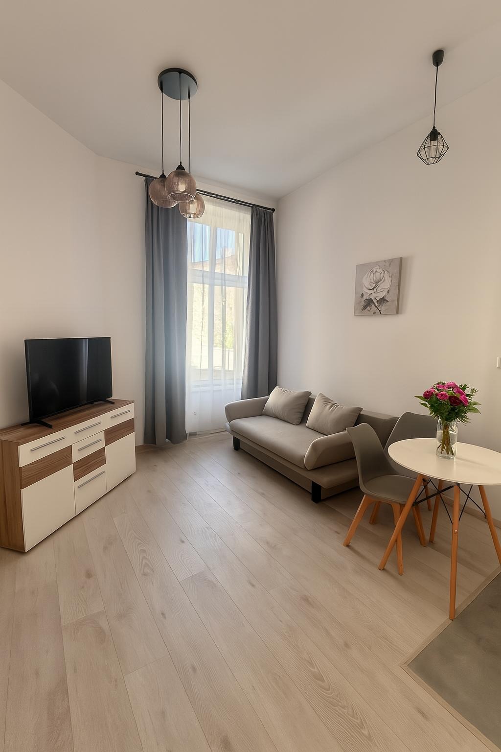 Jasne panele podłogowe w salonie z beżową sofą, stolikiem z kwiatami i komodą pod telewizor. Szare zasłony w oknie i lampy sufitowe.