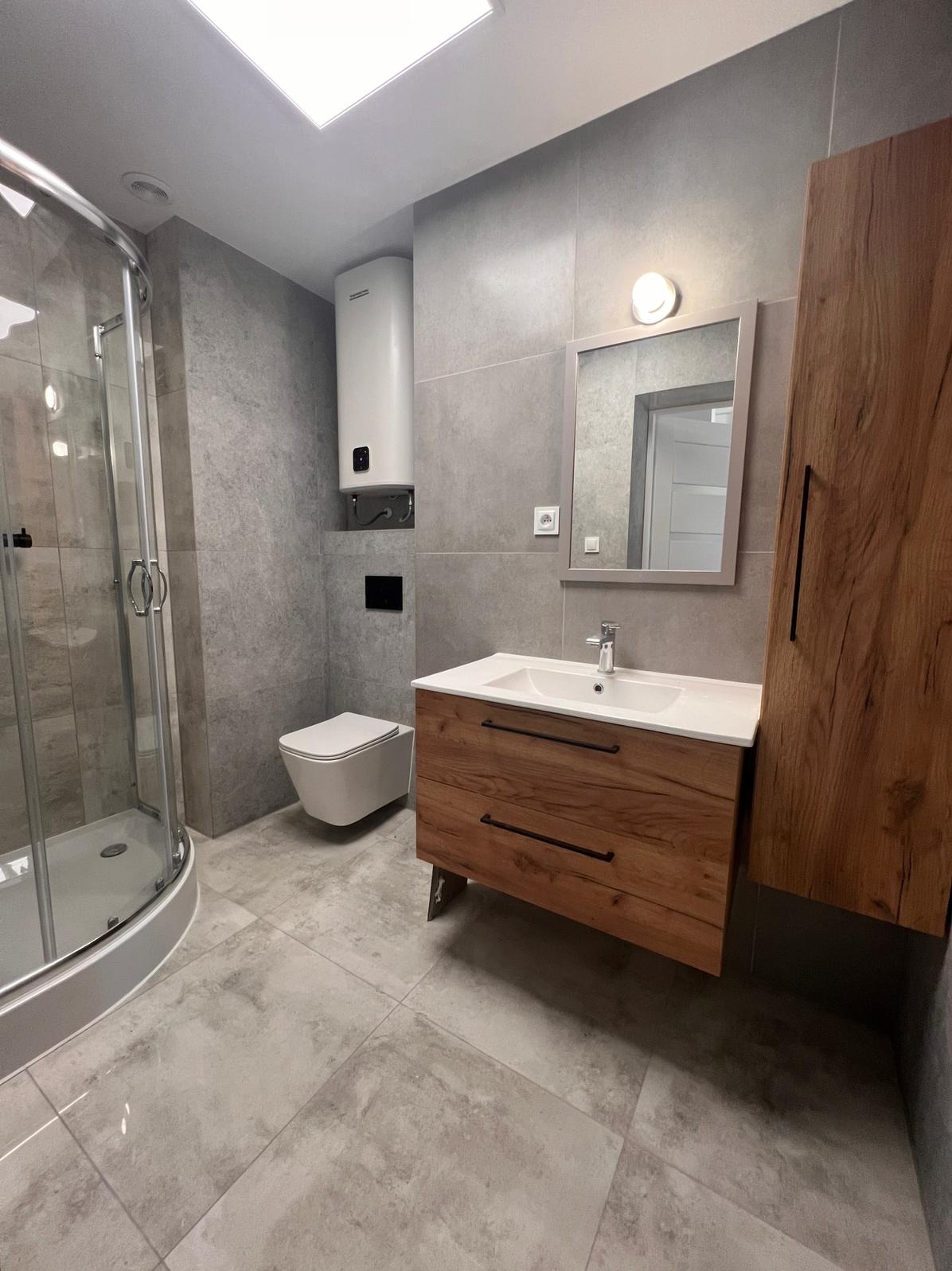 Wykończona łazienka z kabiną prysznicową, umywalką z drewnianą szafką i toaletą w odcieniach szarości z akcentami drewna. Grzejnik elektryczny na ścianie.