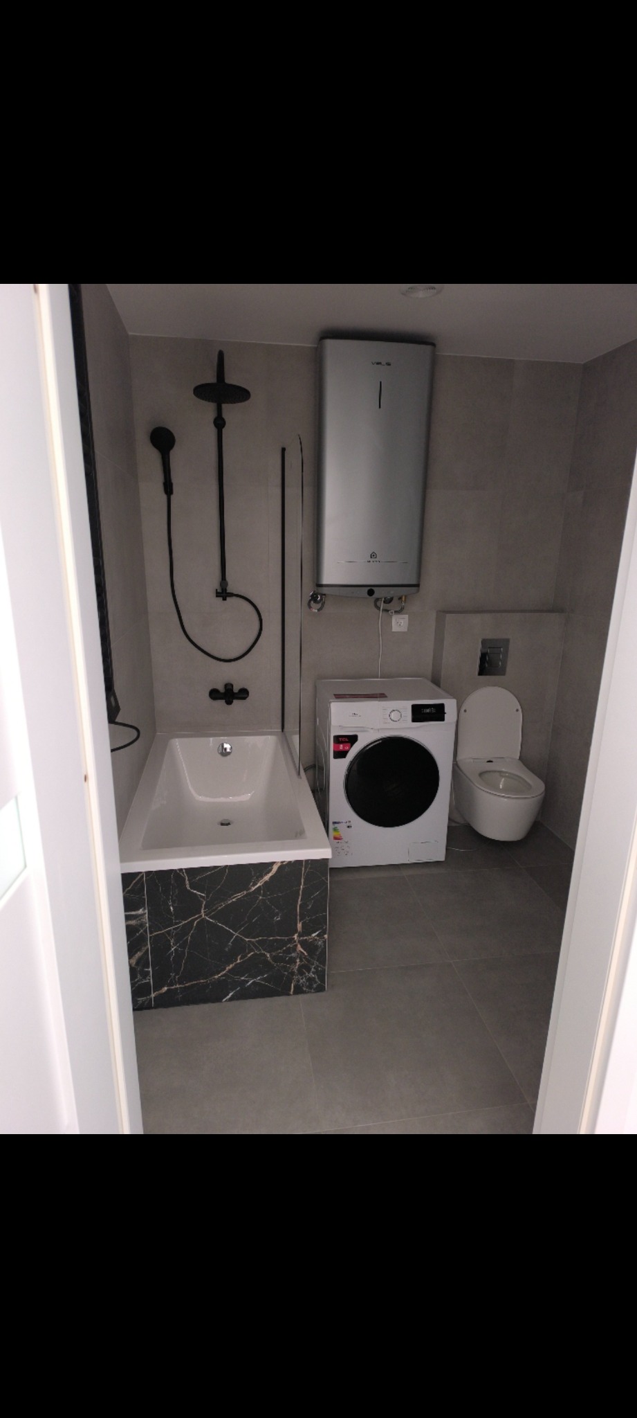 Wyremontowana łazienka w szarościach z wanną obudowaną czarnym marmurem, pralką i bojlerem. Czarna armatura, minimalistyczny design.