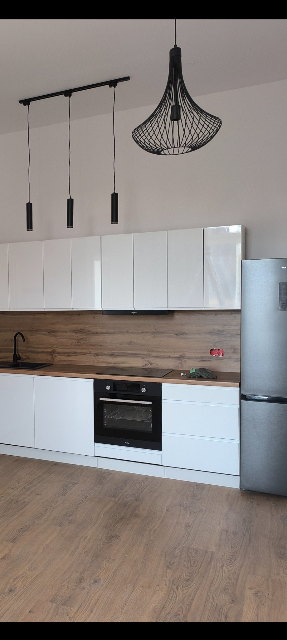 Nowoczesna kuchnia z białymi szafkami, drewnianym blatem i czarnymi lampami wiszącymi. Widoczny sprzęt AGD i panele podłogowe.