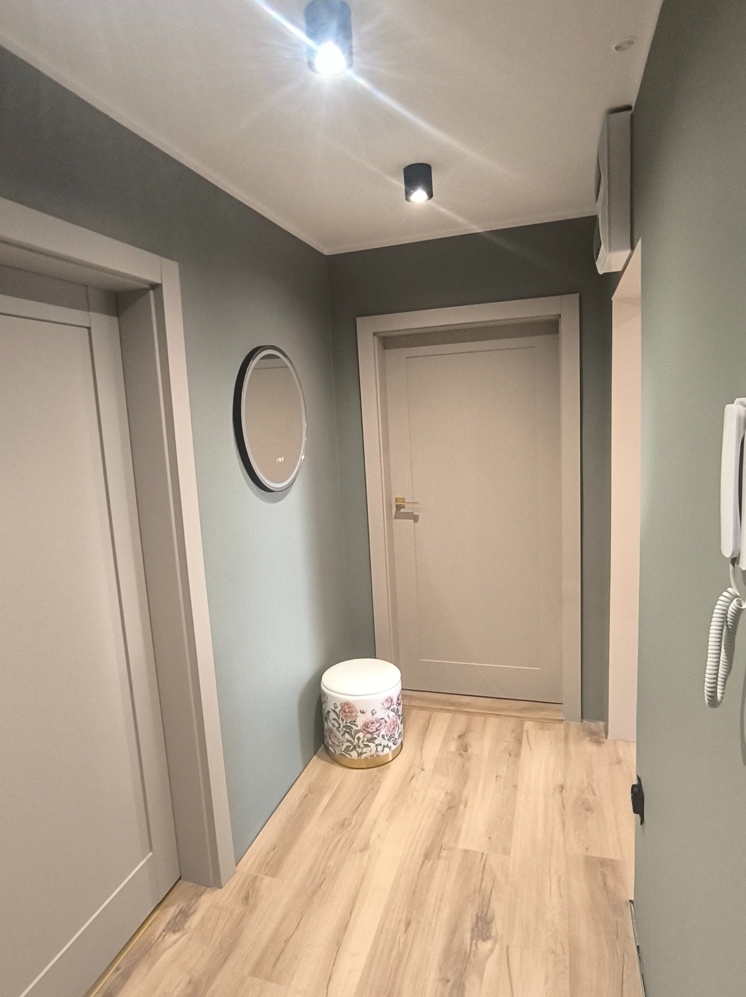 Nowoczesny przedpokój w odcieniach szarości z drewnianą podłogą, okrągłym lustrem i dekoracyjnym stołkiem. Minimalistyczne oświetlenie sufitowe.