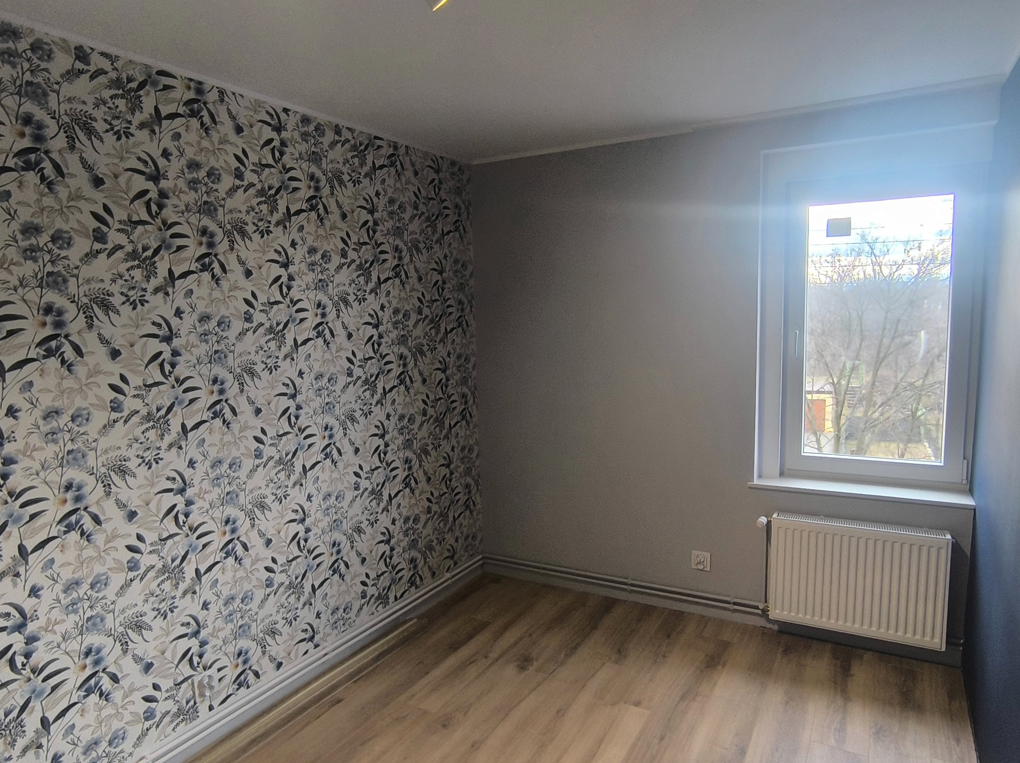 Pokój po remoncie: ściana w kwiatowe wzory, szara ściana, drewniana podłoga, biały grzejnik pod oknem. Widok na zewnątrz przez okno.