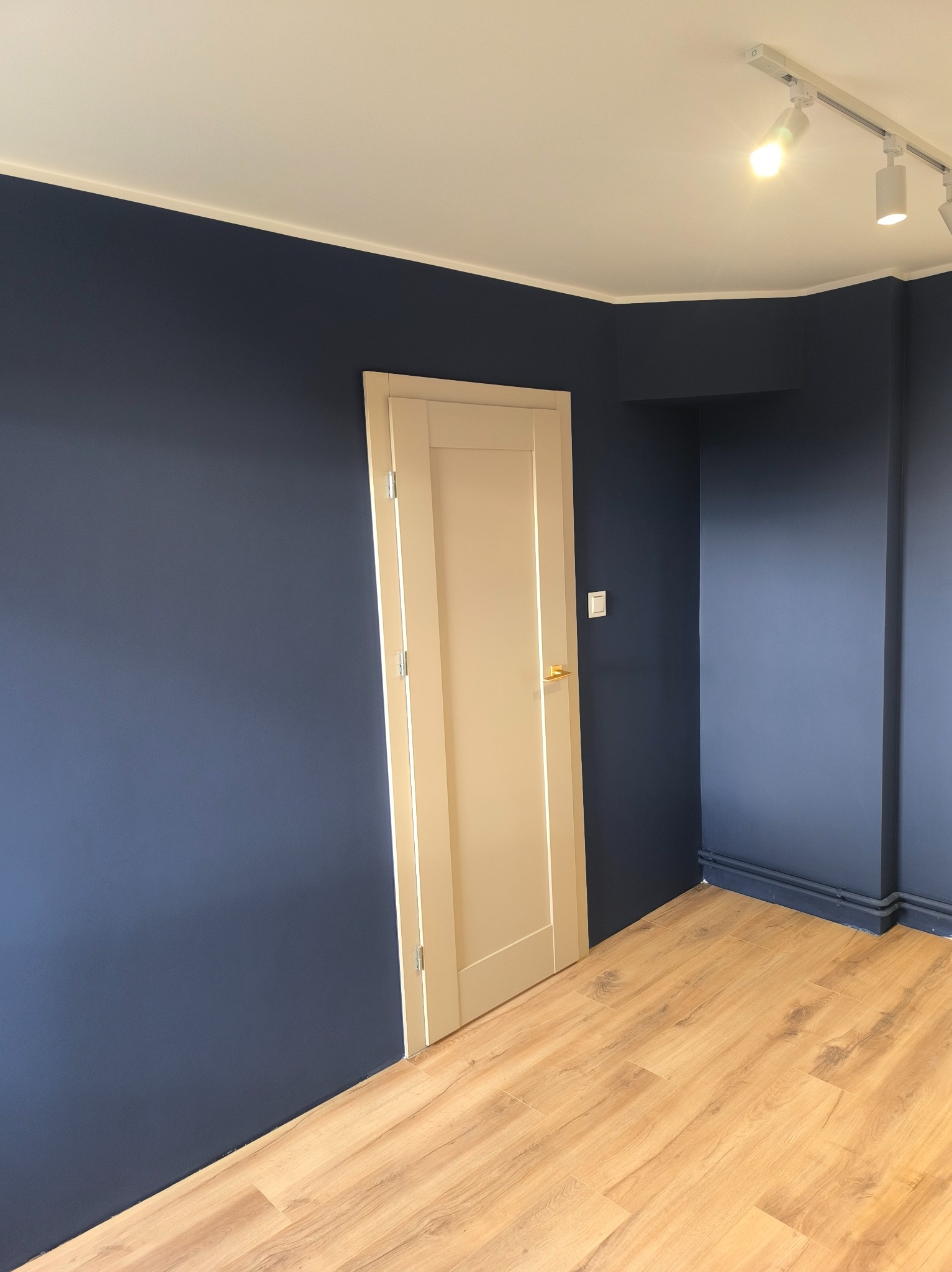 Pokój po malowaniu ścian na granatowo i ułożeniu paneli podłogowych. Widoczne białe drzwi i oświetlenie sufitowe.