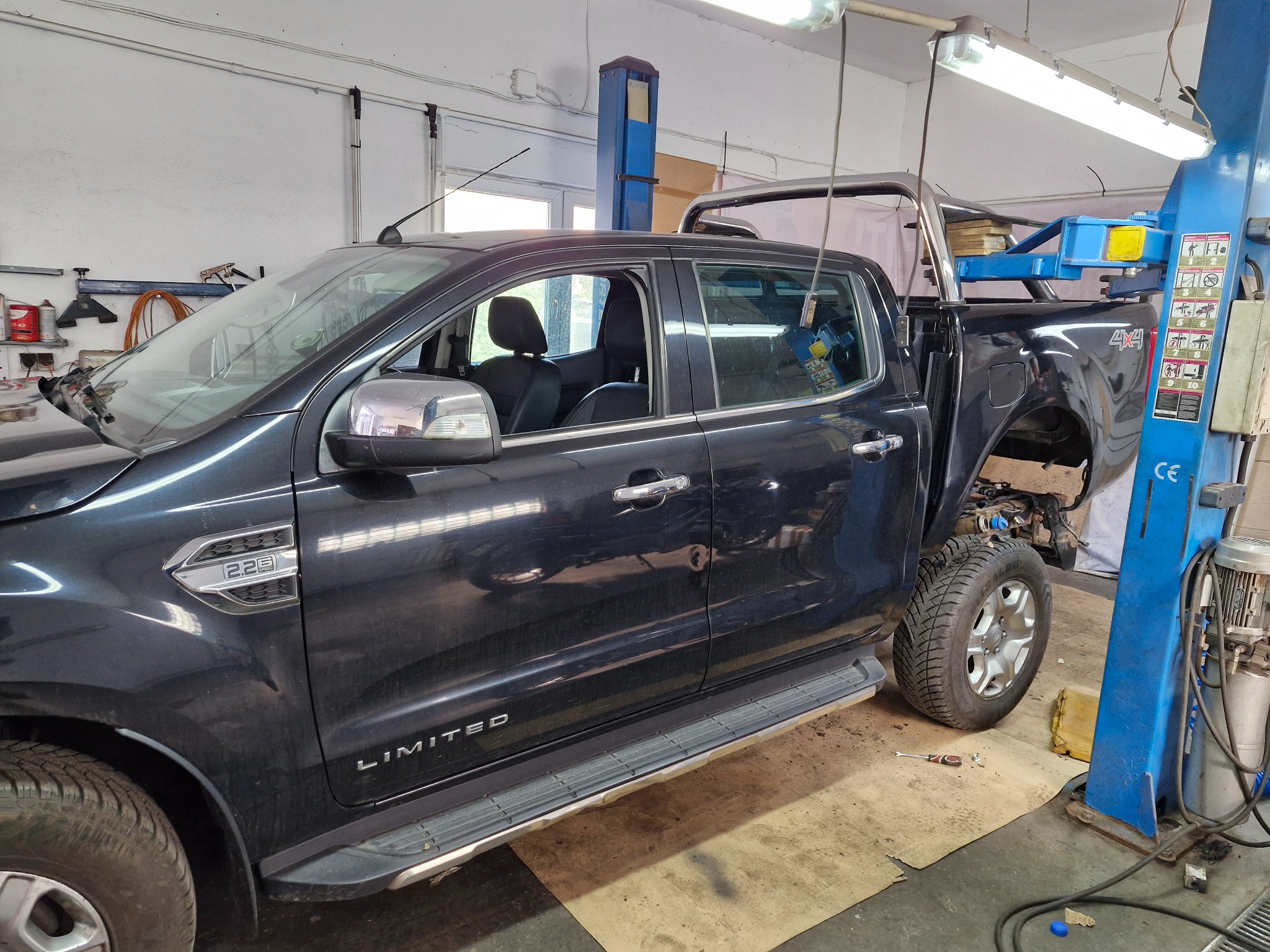Czarny pickup Ford Ranger Limited na podnośniku w warsztacie, z częściowo zdemontowanym nadwoziem, widoczna opona i narzędzie leżące na podłodze.