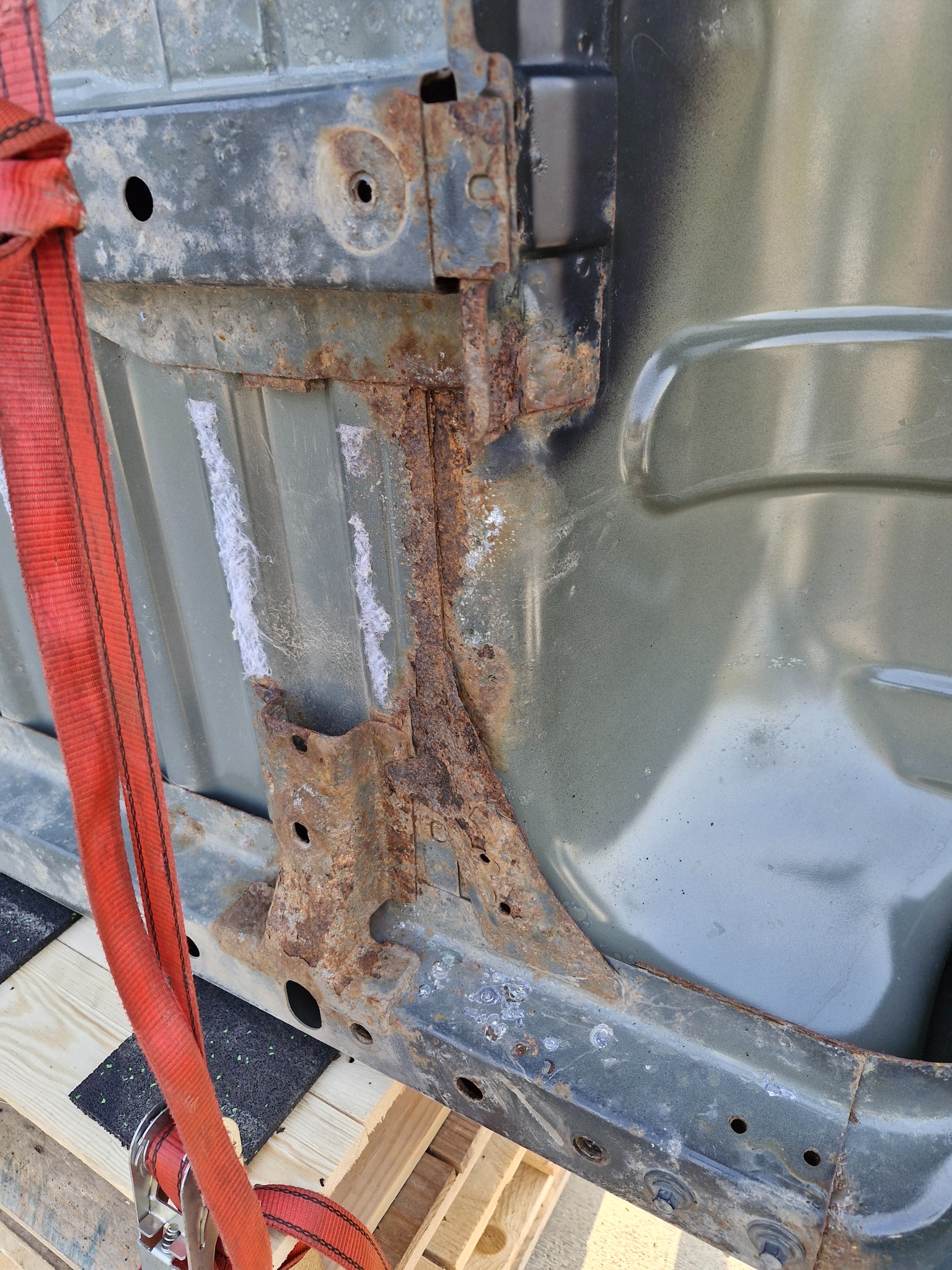 Podwozie samochodu z widoczną korozją na metalowych elementach, zabezpieczone czerwoną taśmą transportową i podłożone na drewnianej palecie.