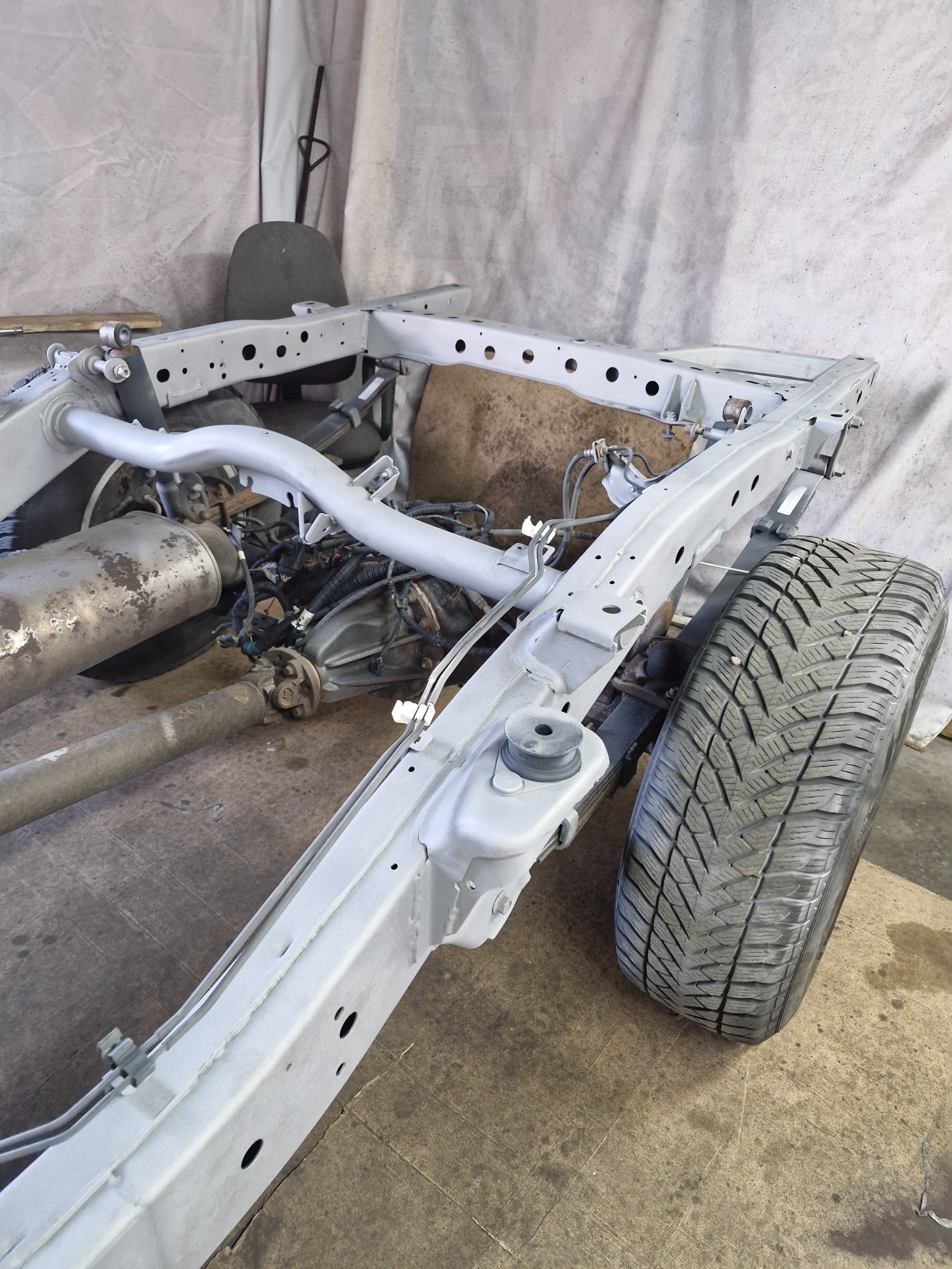 Rama samochodu po piaskowaniu i malowaniu proszkowym, widoczne elementy zawieszenia, silnik i opona zimowa na tle warsztatu.