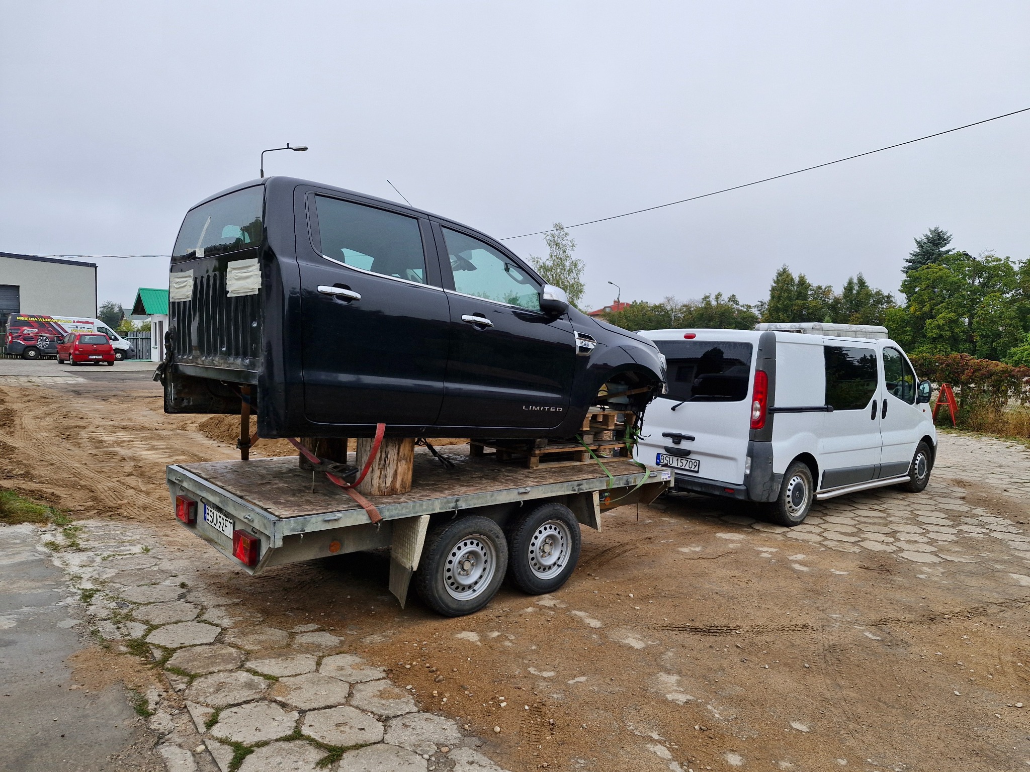 Część karoserii czarnego pickupa, bez kół, transportowana na przyczepie, zaparkowana obok białego busa na placu z betonową kostką.