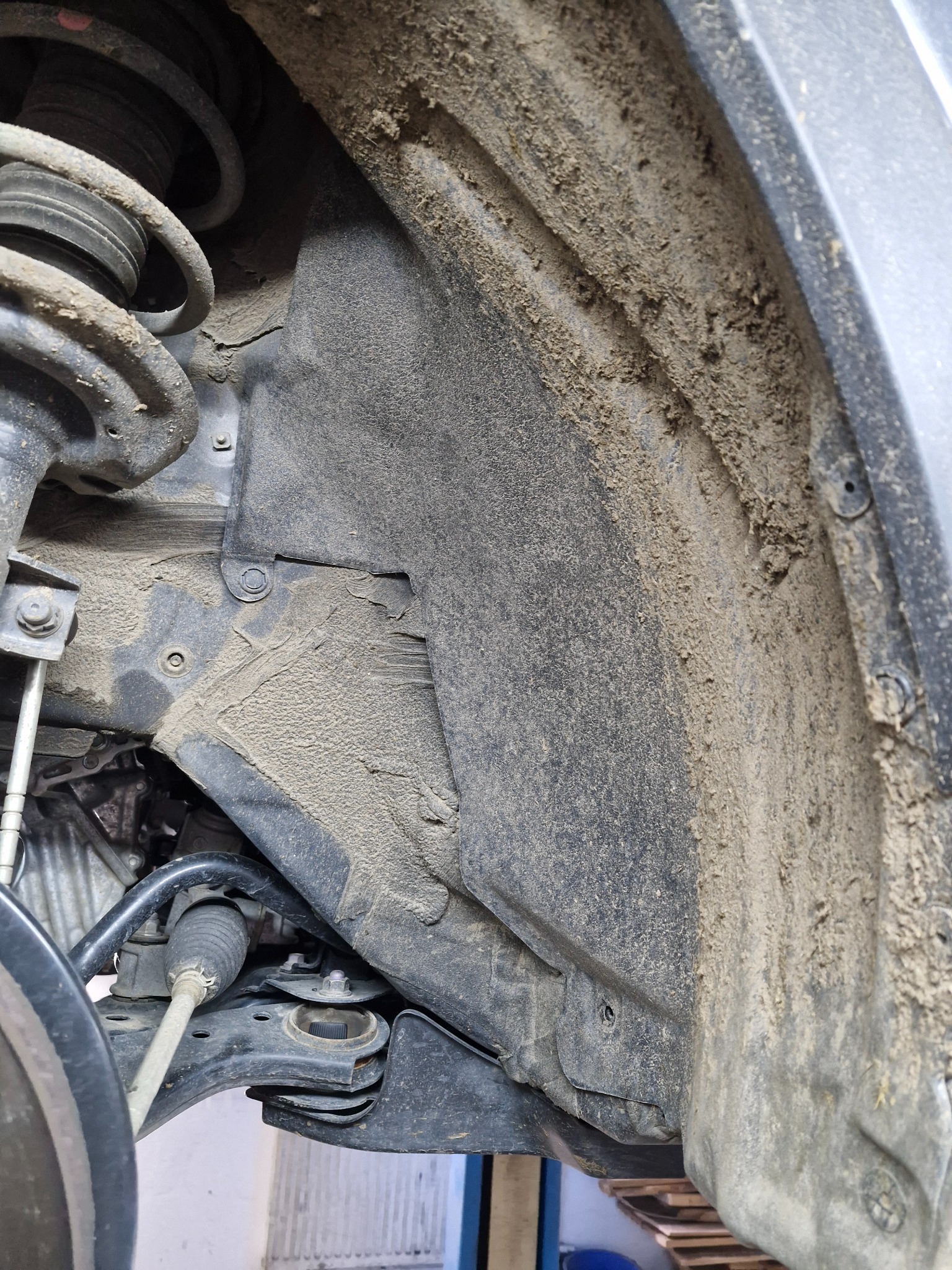 Błotnik i zawieszenie samochodu pokryte grubą warstwą błota, widoczne elementy mechaniczne i mocowania.