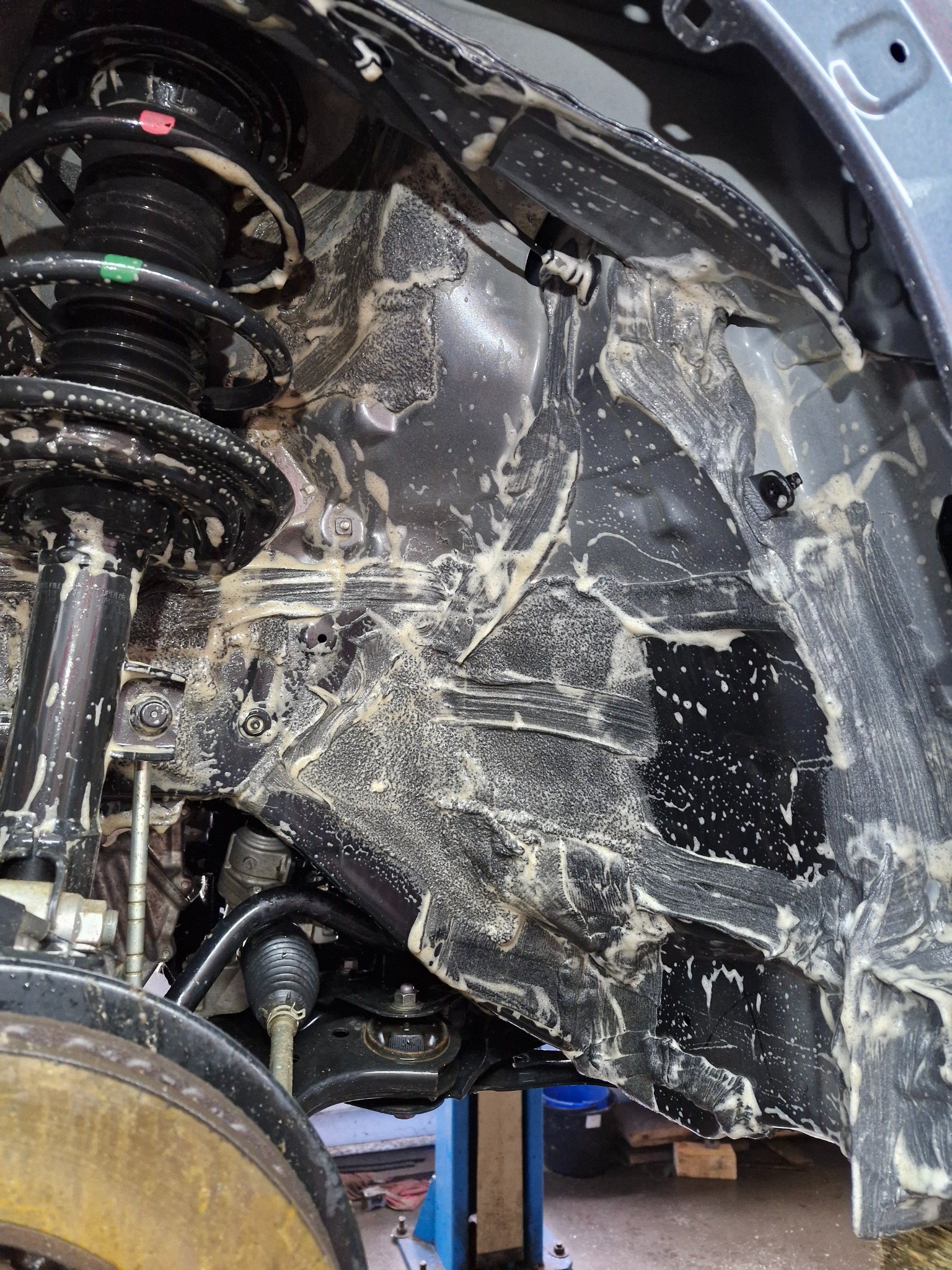 Podwozie samochodu po czyszczeniu, widoczne elementy zawieszenia, amortyzator i nadkole pokryte resztkami środka czyszczącego, w tle niebieski podnośnik warsztatowy.