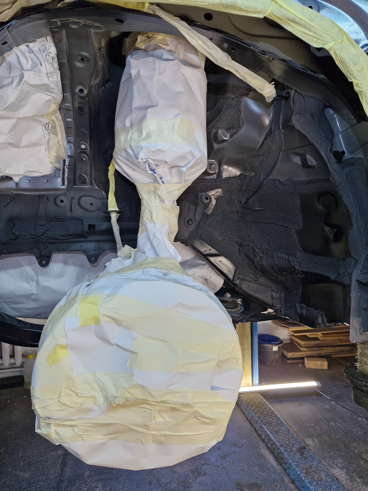 Podwozie samochodu po nałożeniu warstwy ochronnej, zabezpieczone papierem i taśmą malarską przed dalszymi pracami. Widoczne elementy zawieszenia.