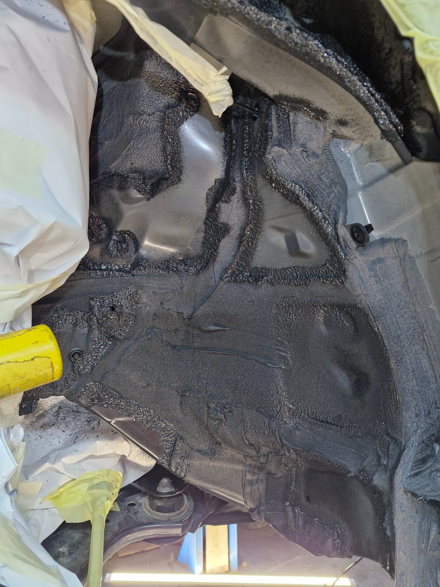 Wnętrze nadkola samochodu po nałożeniu grubej warstwy czarnego preparatu antykorozyjnego, zabezpieczone folią i taśmą malarską.