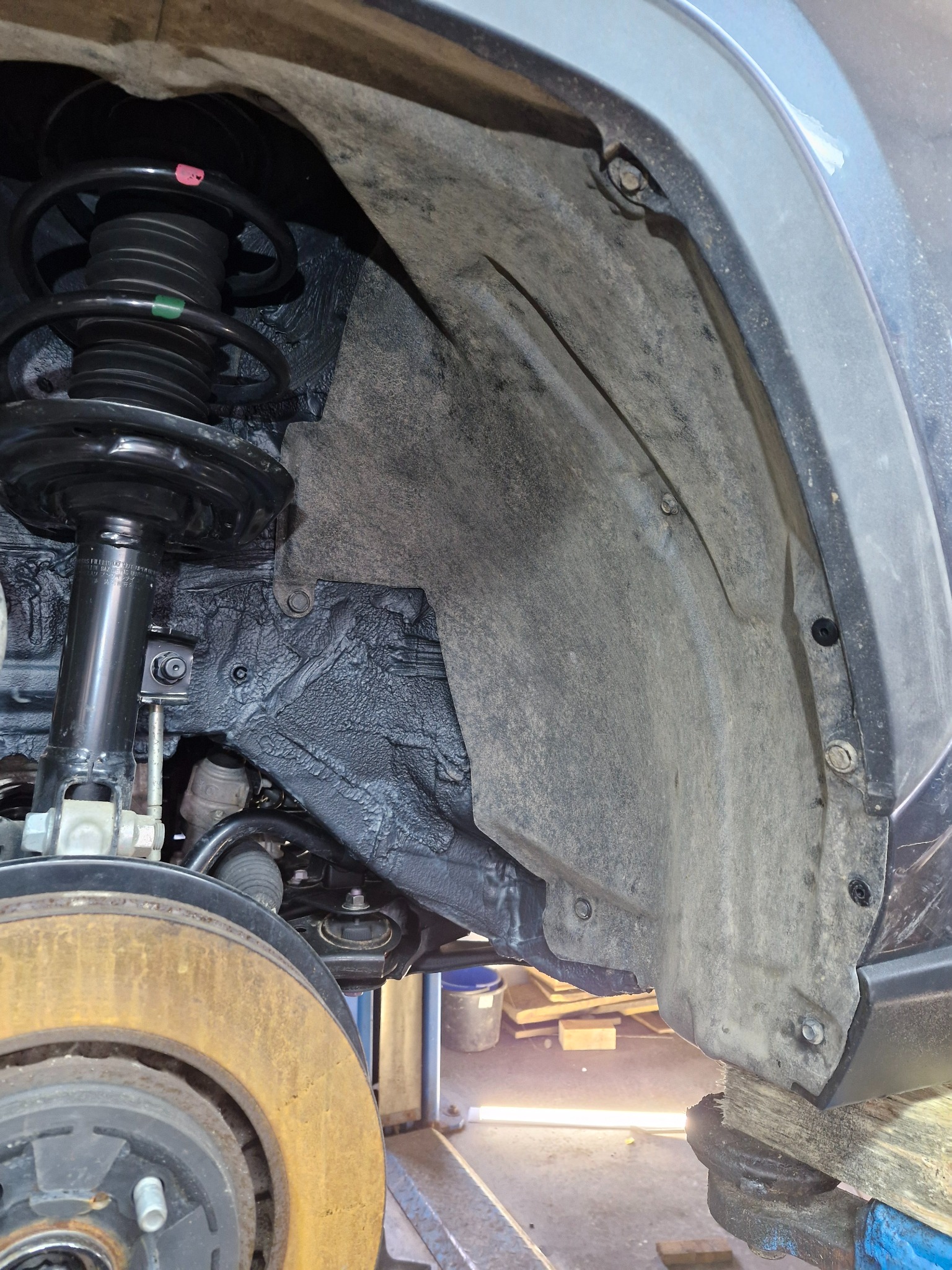 Widok na nadkole samochodu na podnośniku, z widocznym elementem zawieszenia, amortyzatorem, tarczą hamulcową z widocznymi śladami korozji oraz zabezpieczeniem antykorozyjnym na elementach karoserii.