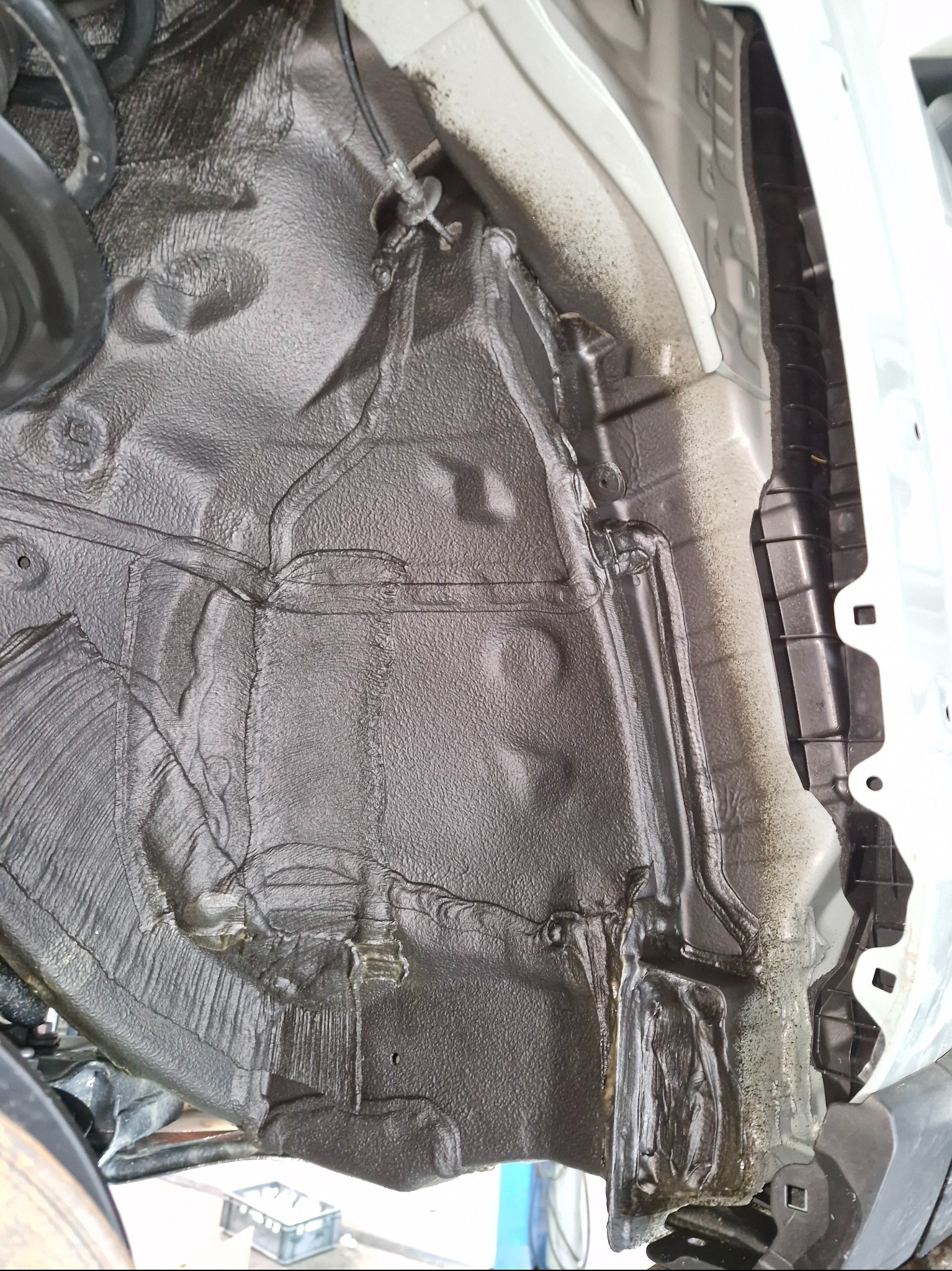 Pokryta preparatem antykorozyjnym podłoga samochodu od strony koła. Widoczne elementy zawieszenia i nadkola. Detal po zabiegu.