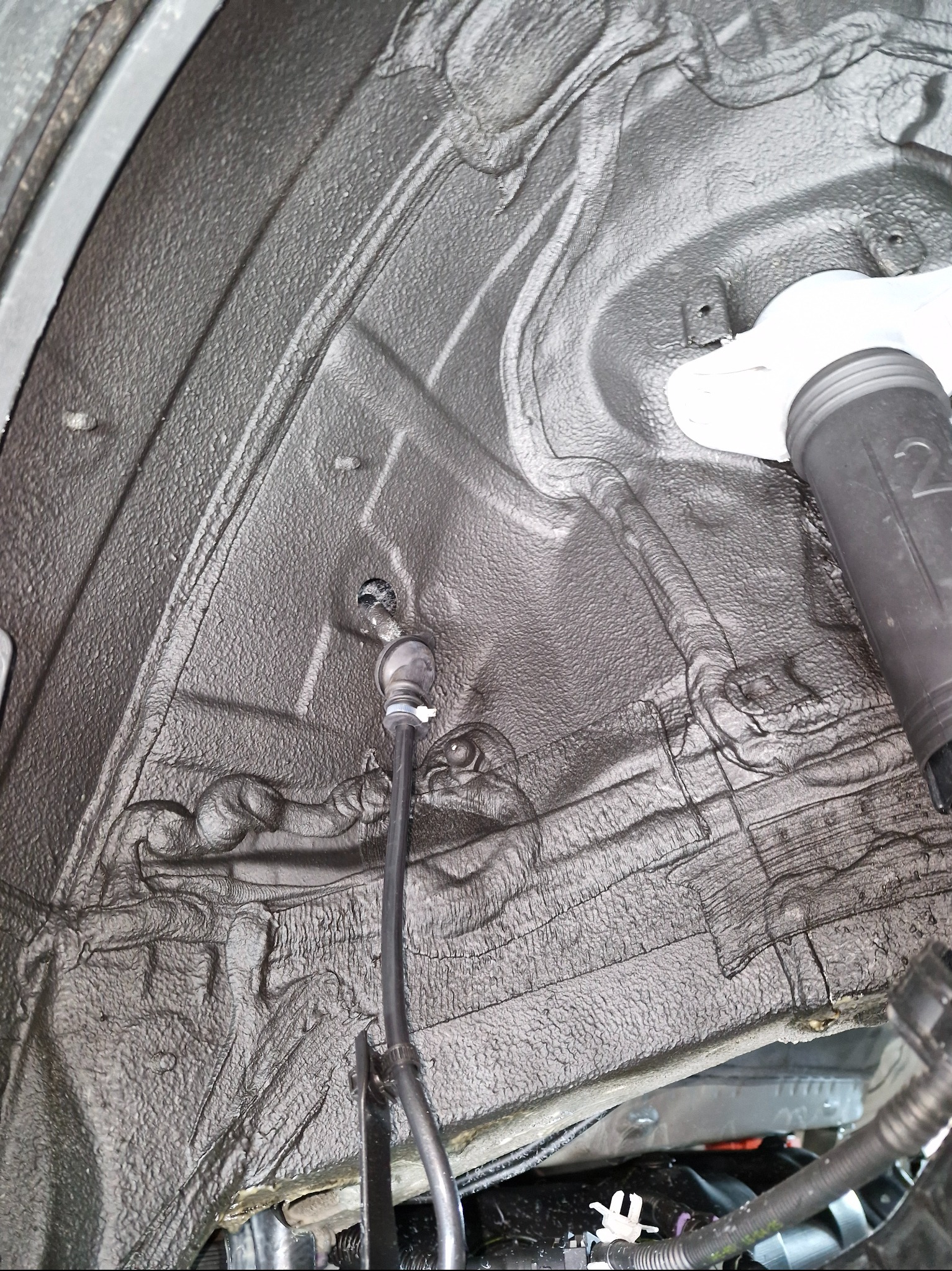 Podwozie auta po konserwacji antykorozyjnej, widoczne elementy zawieszenia i przewody. Szara, chropowata powierzchnia zabezpieczona specjalistycznym preparatem.