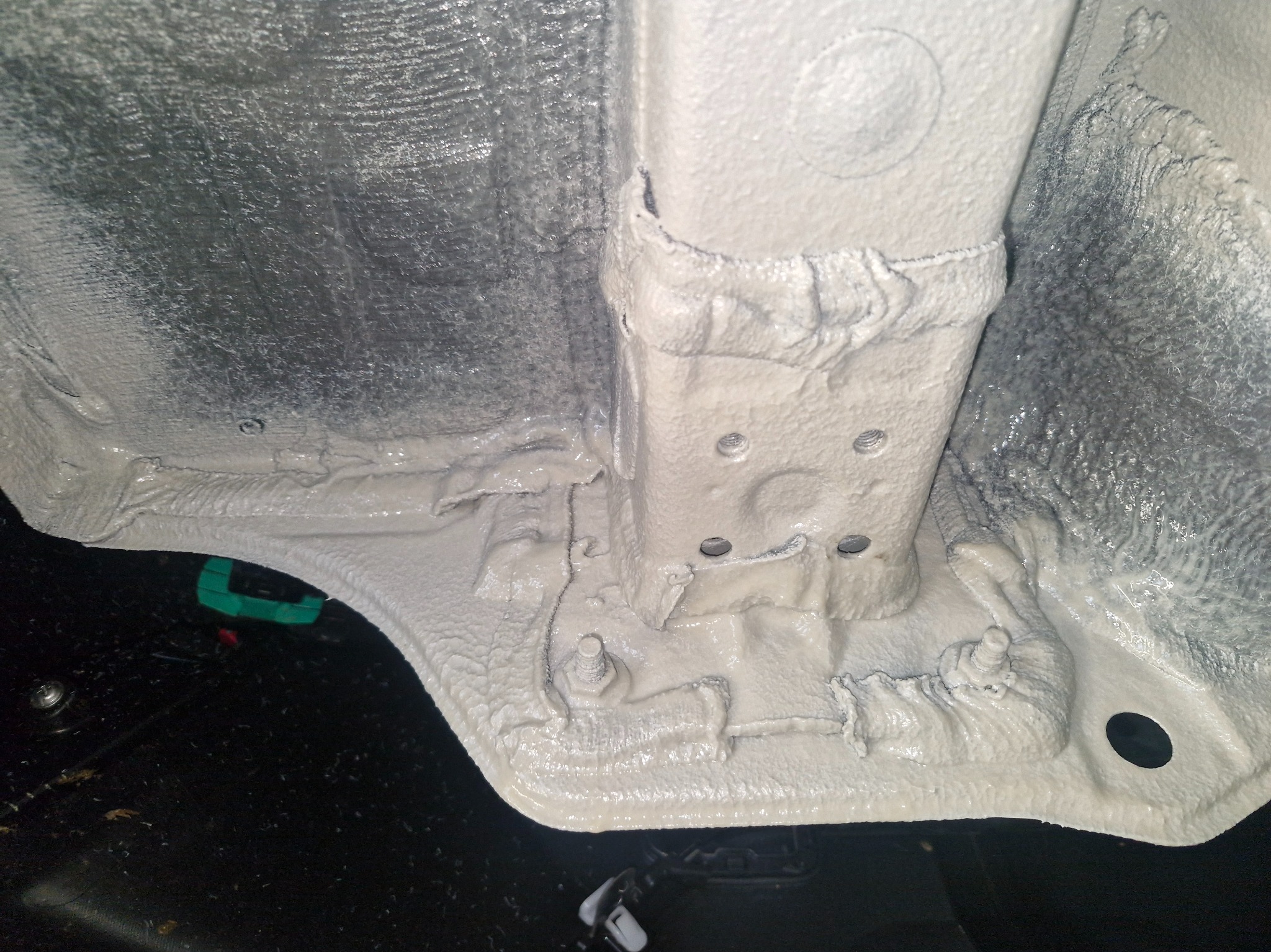 Podwozie auta po nałożeniu ochronnej powłoki. Widoczne elementy konstrukcyjne pokryte gęstą, jasną substancją. Fragment czarnego elementu w dolnej części kadru.