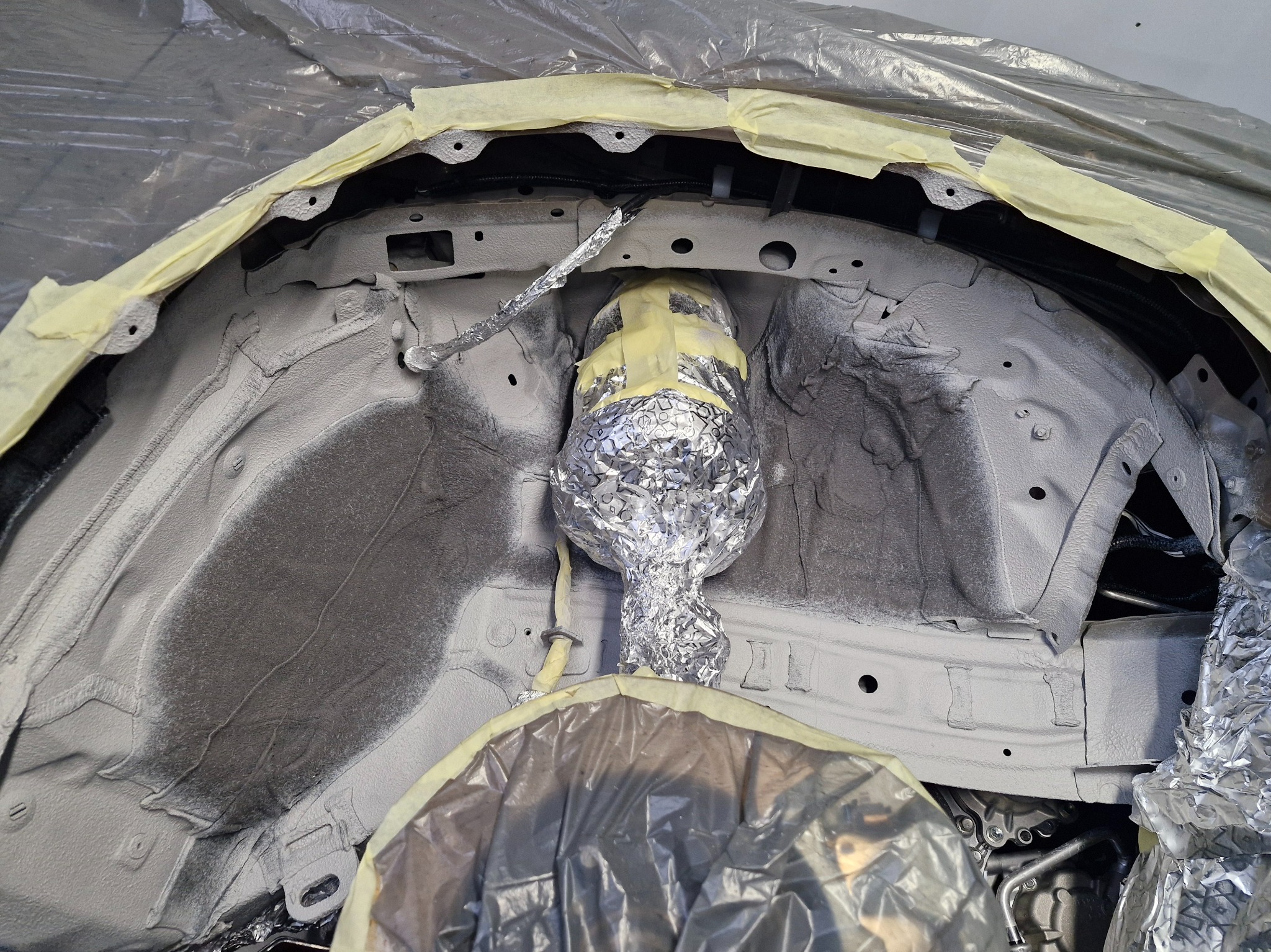 Podwozie auta po nałożeniu szarej warstwy ochronnej, zabezpieczone folią i taśmą. Widoczne elementy zawieszenia owinięte folią aluminiową. Detal konserwacji.