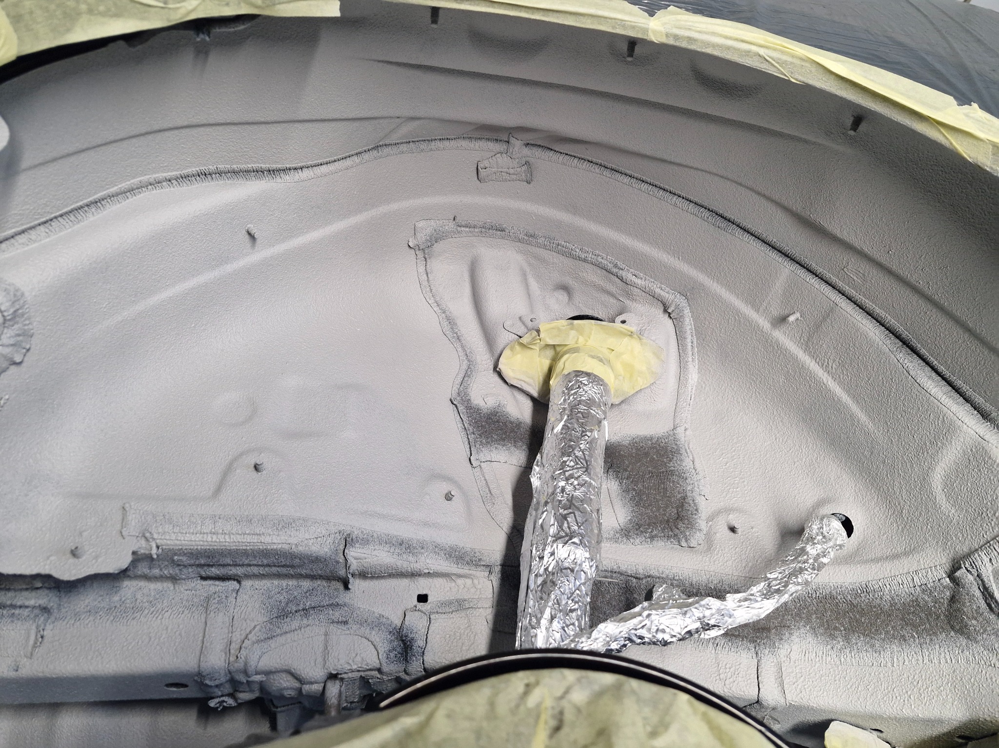 Wnętrze nadkola samochodu po piaskowaniu i nałożeniu warstwy ochronnej, zabezpieczone taśmą i folią aluminiową. Widoczne detale konstrukcyjne.