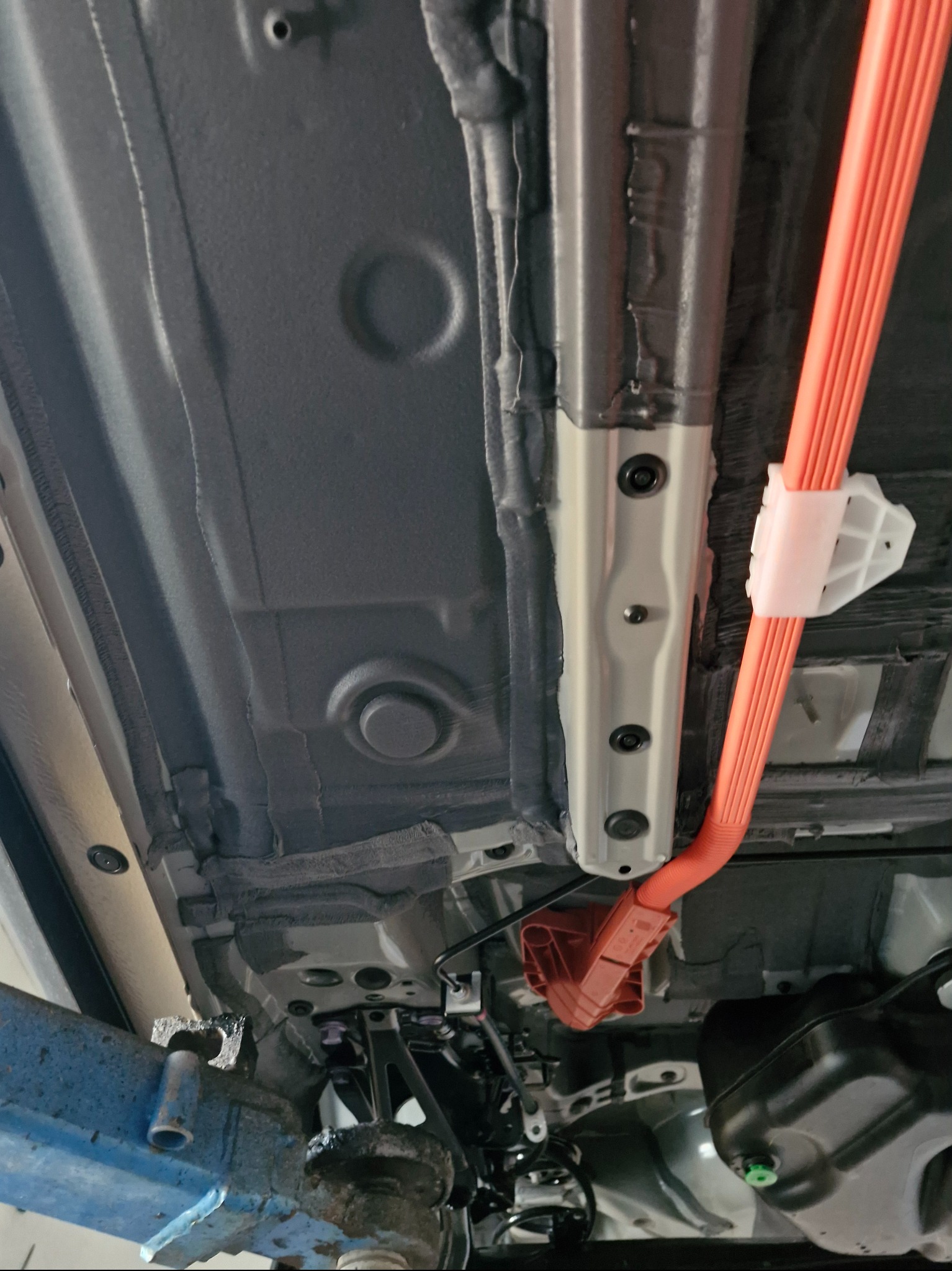 Spód pojazdu po konserwacji. Widoczne zabezpieczenie antykorozyjne, elementy zawieszenia i pomarańczowe przewody. Auto na podnośniku.