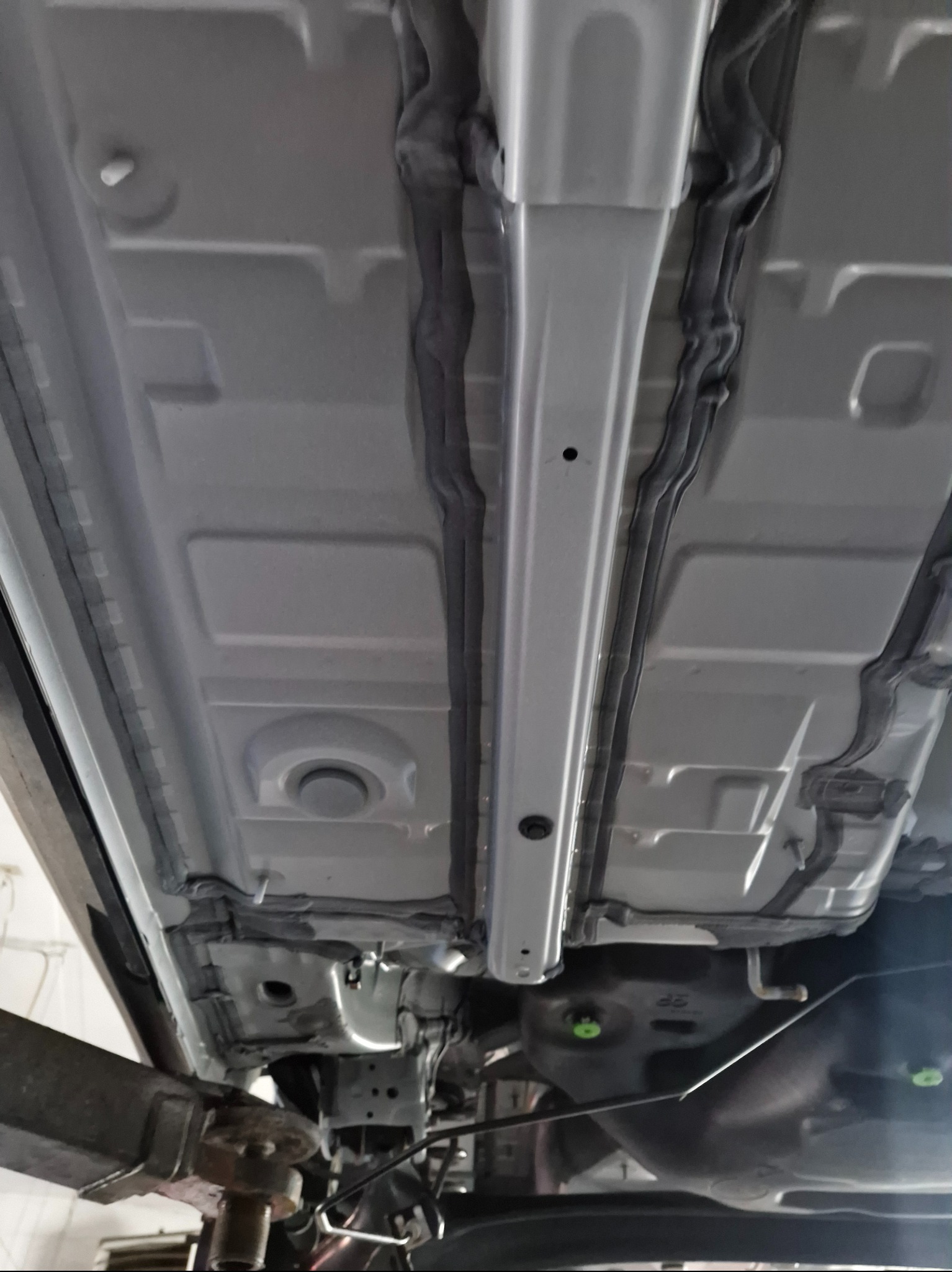 Widok podwozia samochodu na podnośniku, po konserwacji. Widoczne elementy konstrukcyjne i zabezpieczenia antykorozyjne w kolorze szarym i czarnym.