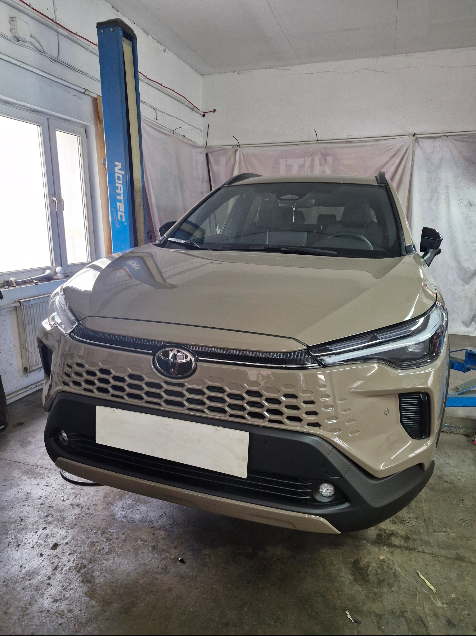 Beżowy Toyota Corolla Cross na podnośniku w warsztacie. Widok z przodu, na pierwszym planie atrapa tablicy rejestracyjnej. W tle okno i niebieski podnośnik.