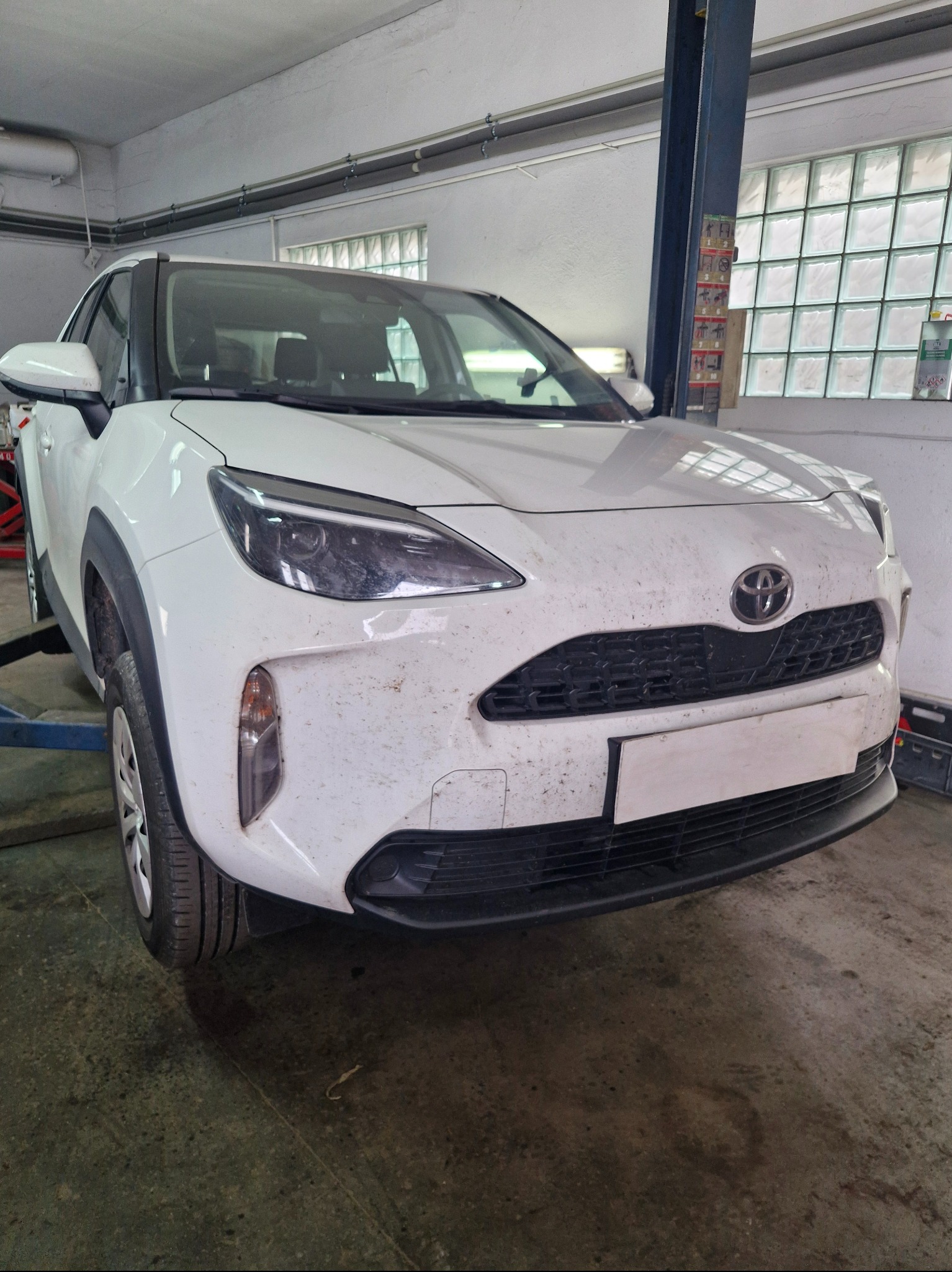 Biała Toyota Yaris Cross na podnośniku w warsztacie, przygotowana do konserwacji. Widoczne ślady zabrudzeń na karoserii.