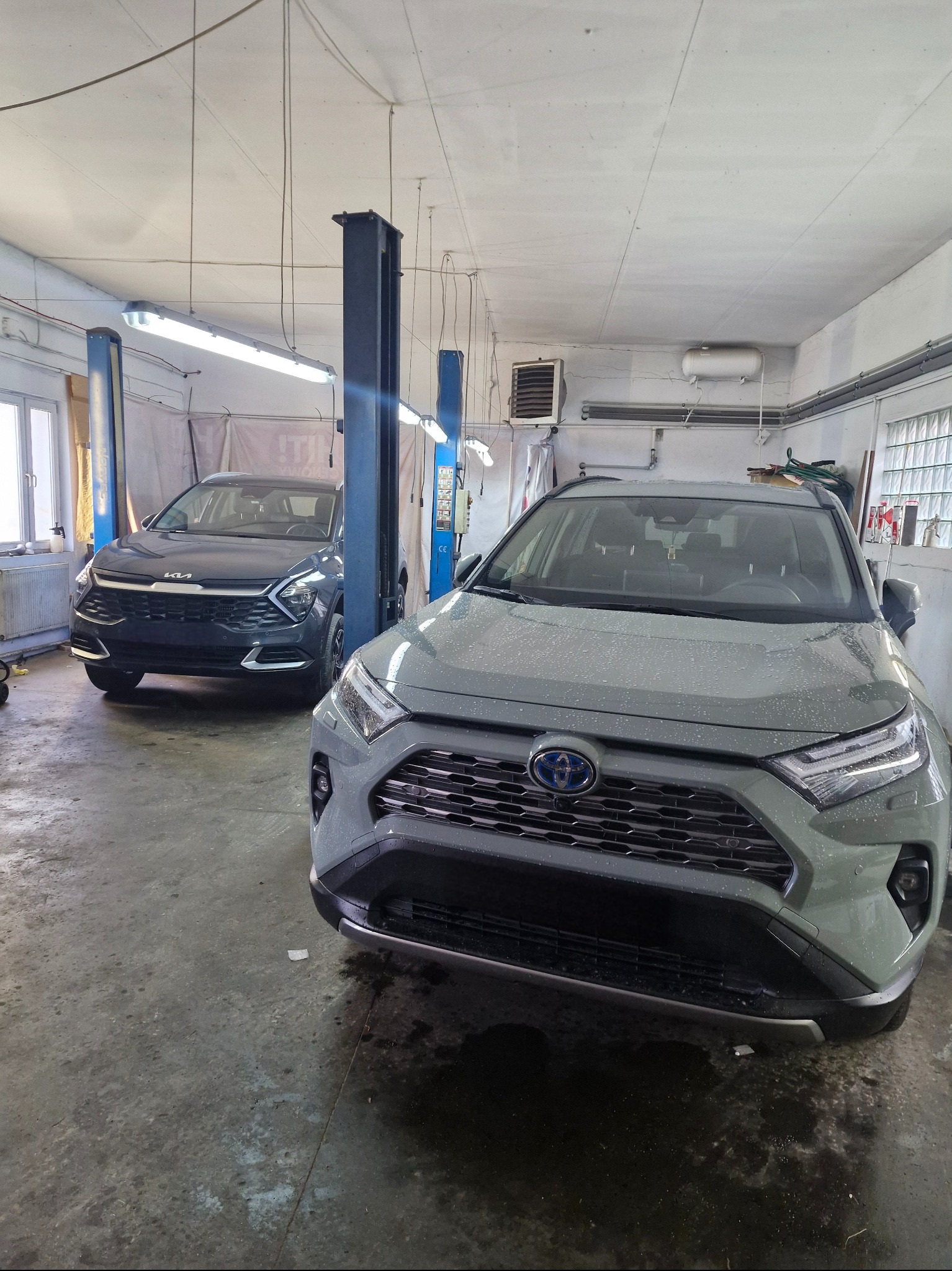 Dwa auta w warsztacie: szary SUV Toyota z przodu, ciemny Kia na podnośniku w tle. Widoczne wyposażenie warsztatu, w tym podnośniki kolumnowe.