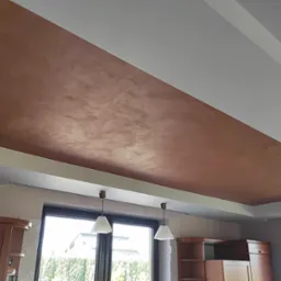 Fragment sufitu z dekoracyjną, brązową fakturą przypominającą trawertyn, oświetlony wiszącymi lampami, widok z dołu, fragment zabudowy kuchennej w tle.