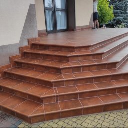 F. U. DUBAJ Joanna Dubaj - Zewnętrzne schody wejściowe wyłożone brązowymi, kwadratowymi płytkami ceramicznymi z jasną fugą, prowadzące do drzwi wejściowych.