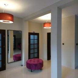 F. U. DUBAJ Joanna Dubaj - Jasne, przestronne wnętrze z beżowymi ścianami, białymi kolumnami i błyszczącą, kremową podłogą. Widoczne dwa ciemne drzwi z przeszkleniami, duże lustro odbijające fioletowy puf i lampę z abażurem...