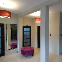 F. U. DUBAJ Joanna Dubaj - Jasne wnętrze korytarza z kremowymi ścianami, białymi płytkami podłogowymi, ciemnymi drzwiami z szybkami, dużym lustrem i fioletowym pufem z guzikami, oświetlone wiszącymi lampami z abażurami...