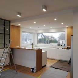 F. U. DUBAJ Joanna Dubaj - Wnętrze kuchni w trakcie wykańczania, widoczna aluminiowa drabina, zabezpieczona podłoga, białe szafki kuchenne z drewnianymi elementami, okno z widokiem na dom.