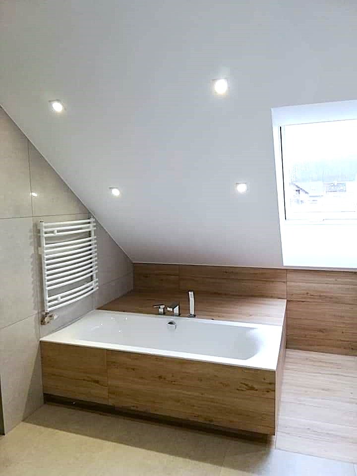 Biała wanna obudowana panelami imitującymi drewno w łazience na poddaszu z oknem dachowym i grzejnikiem drabinkowym