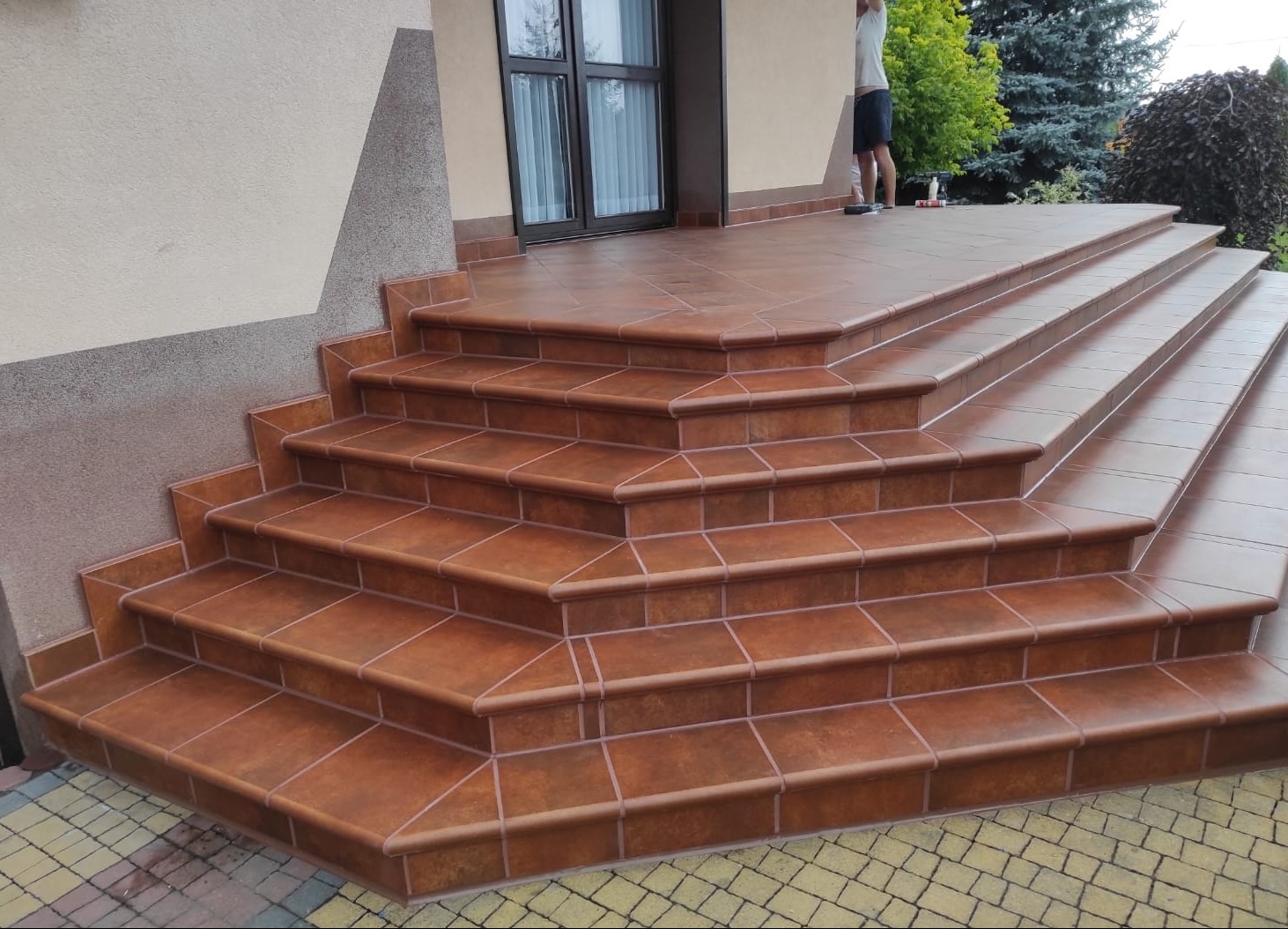 Zewnętrzne schody wejściowe wyłożone brązowymi, kwadratowymi płytkami ceramicznymi z jasną fugą, prowadzące do drzwi wejściowych.