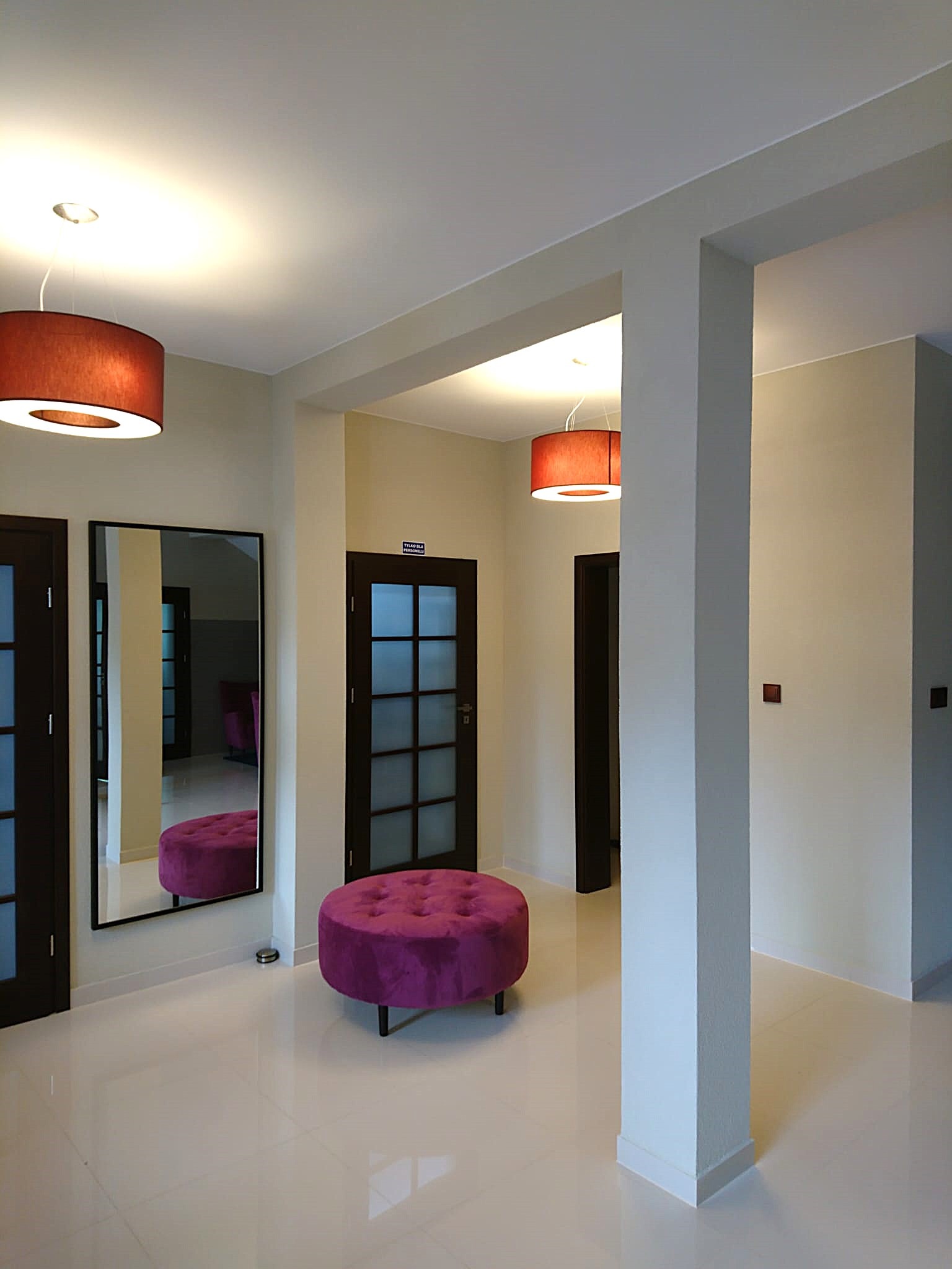 Jasne wnętrze korytarza z kremowymi ścianami, białymi płytkami podłogowymi, ciemnymi drzwiami z szybkami, dużym lustrem i fioletowym pufem z guzikami, oświetlone wiszącymi lampami z abażurami...