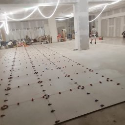 WEBEKO Maciej Paśnik - Przygotowanie do układania płytek w dużym pomieszczeniu, widoczne poziomowanie płytek za pomocą klipsów i klinów, w tle oświetlenie budowlane i pracownicy.