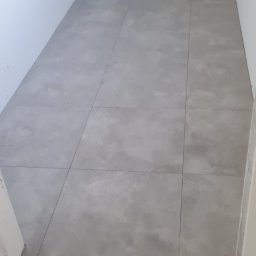 WEBEKO Maciej Paśnik - Szare, matowe płytki podłogowe ułożone w pomieszczeniu w trakcie prac wykończeniowych, widoczne niedoskonałości na ścianach i brak listew przypodłogowych.